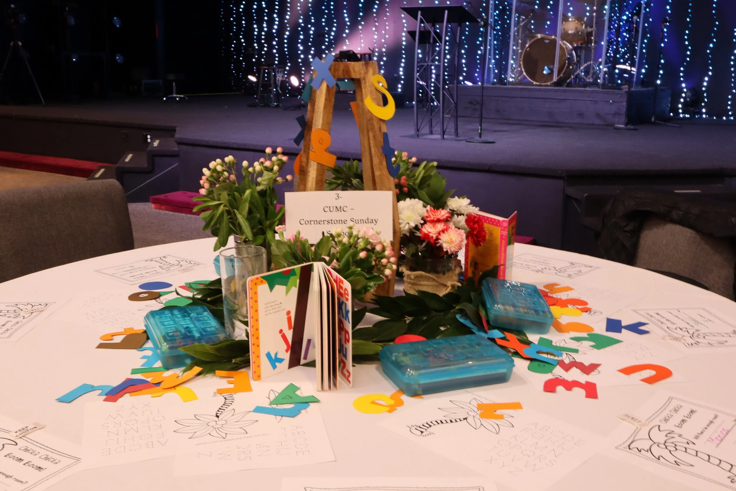 A round table decorated with flowers, coloring pages, and small plastic containers, with a stage in the background decorated with string lights.