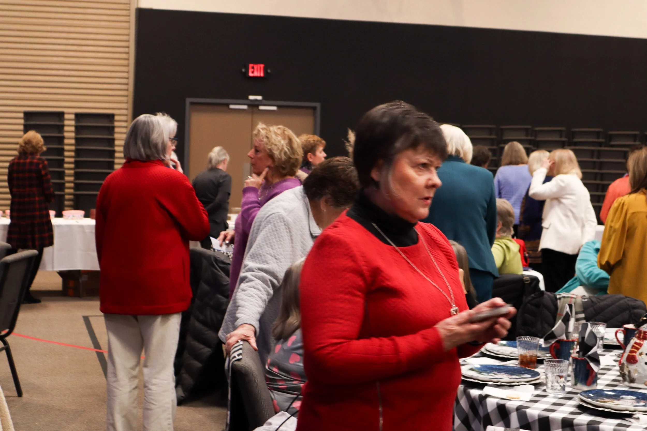 People gathered at a social event, standing around tables with plates, drinks, and tableware, in a room with dark walls and a wood-paneled wall.