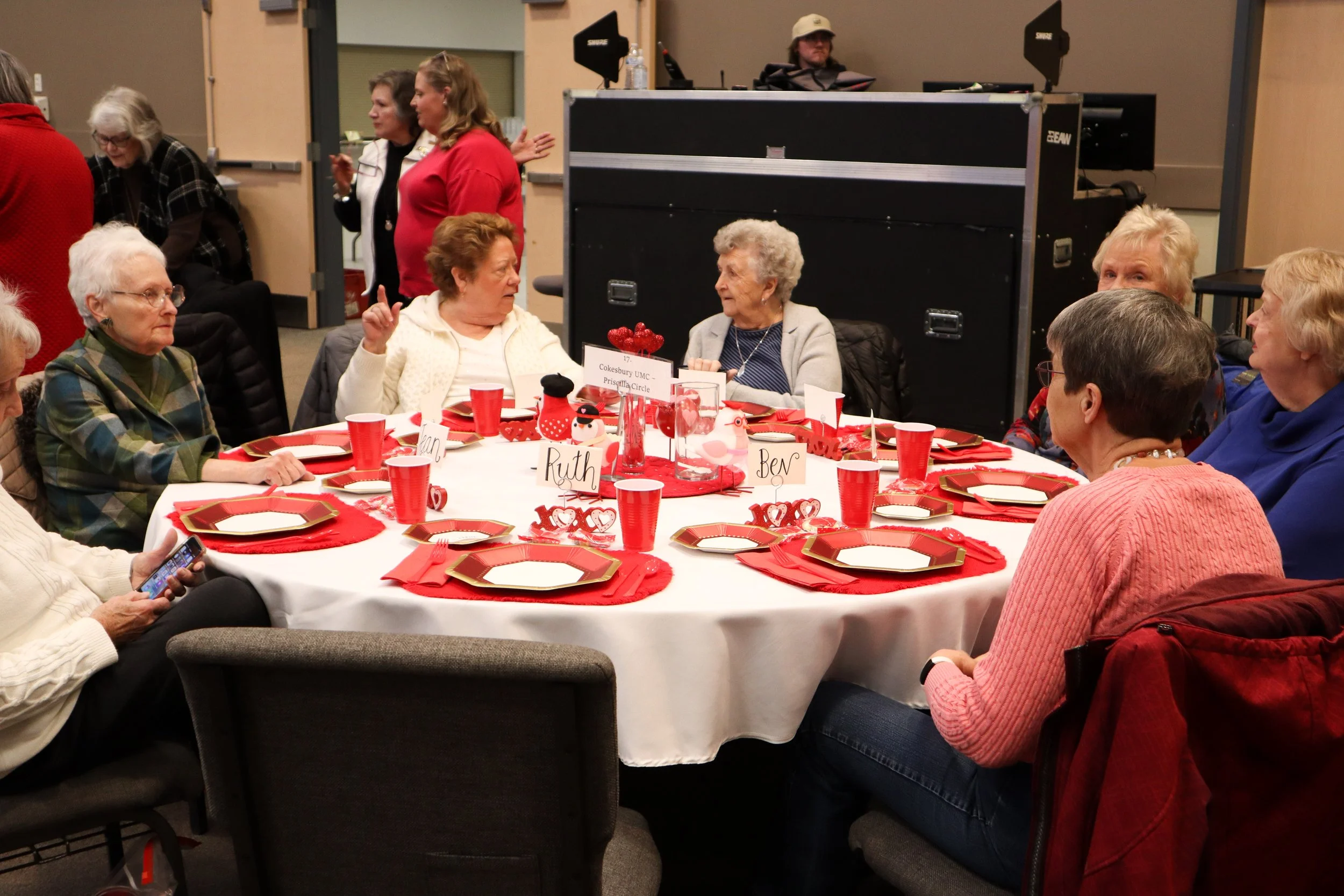 Group of elderly women sitting around a round table decorated with red and white tableware for a holiday celebration, with some women engaged in conversation and others using their phones. In the background, a DJ is standing behind equipment as other