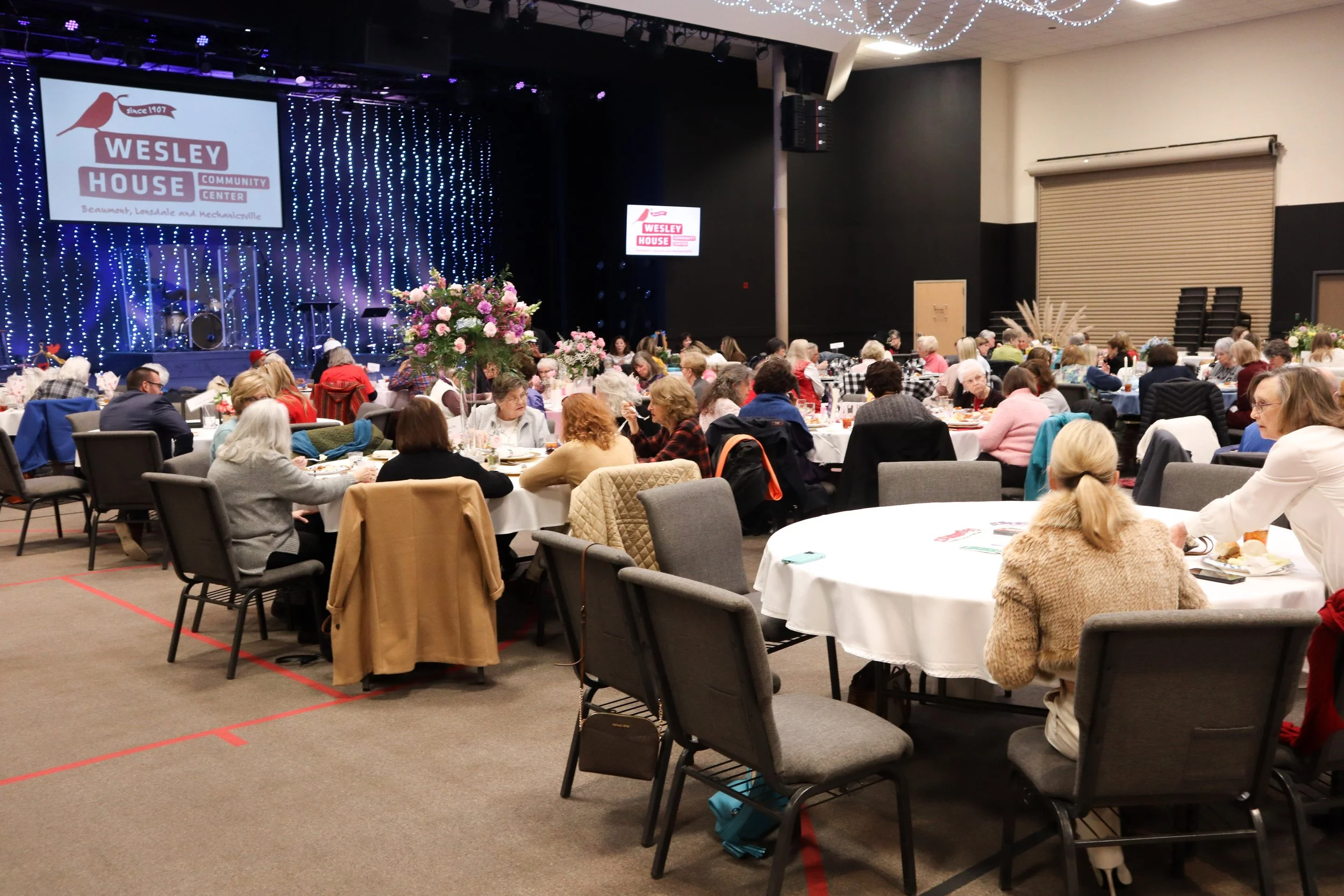 A large indoor gathering at Wesley House Community Center with multiple round tables set for a meal, surrounded by mostly elderly people. The stage features blue string lights, and there are two screens displaying the Wesley House logo.