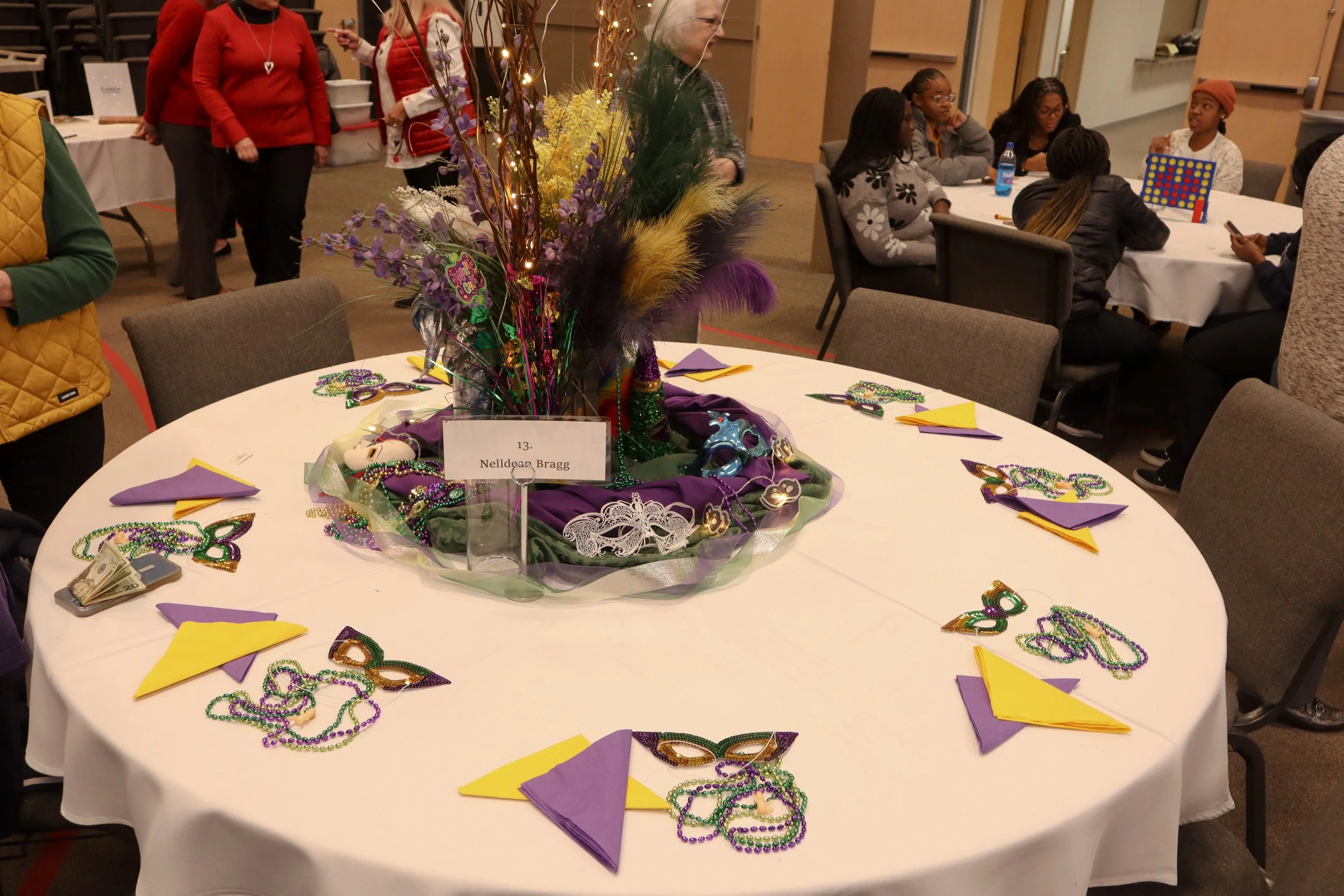 Decorated Mardi Gras-themed table with masks, beads, and colorful napkins in purple and yellow, surrounded by people at a festive gathering.
