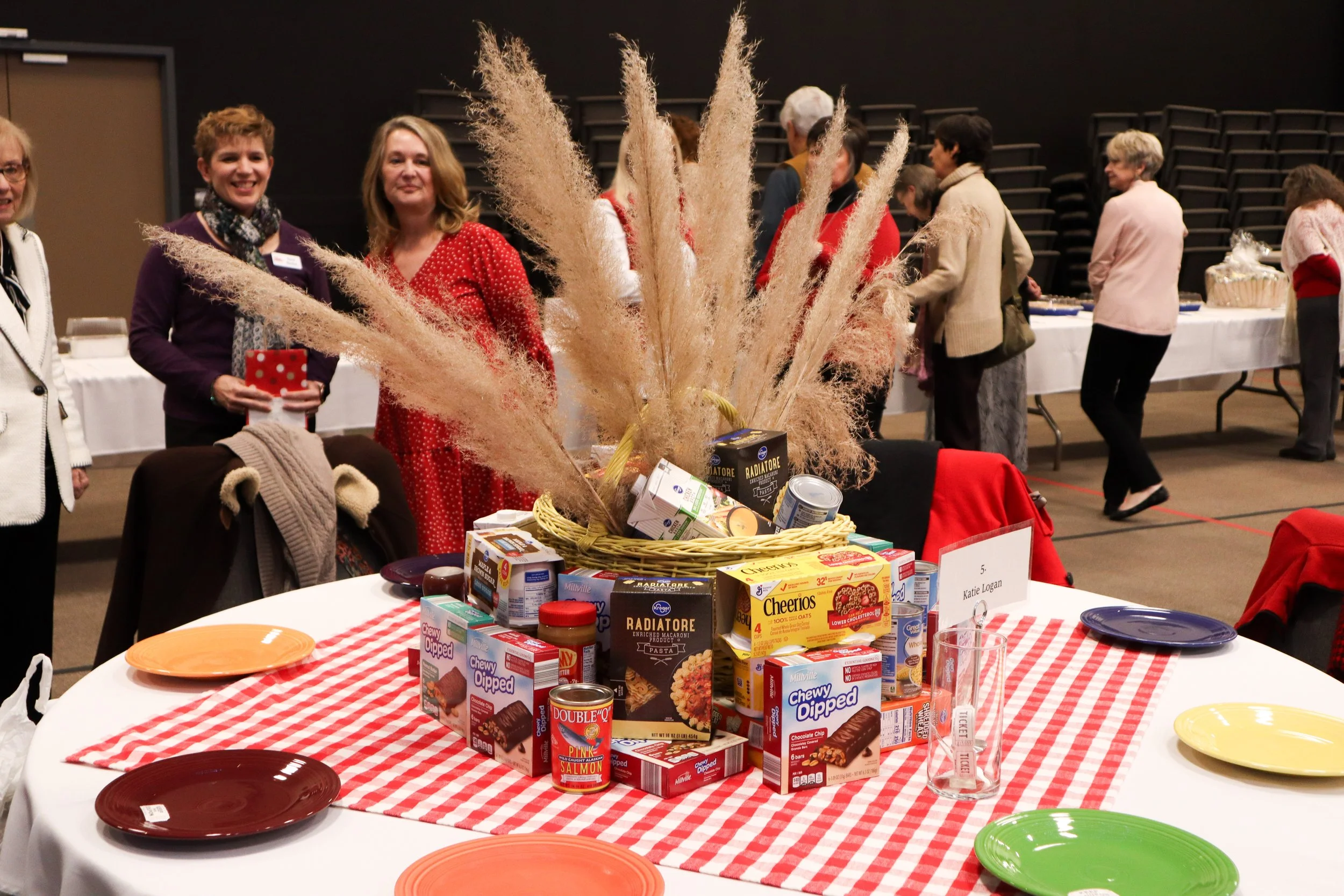 A holiday food drive table with canned goods, boxed food, and a decorative centerpiece of pampas grass, surrounded by people in festive attire at a community event.