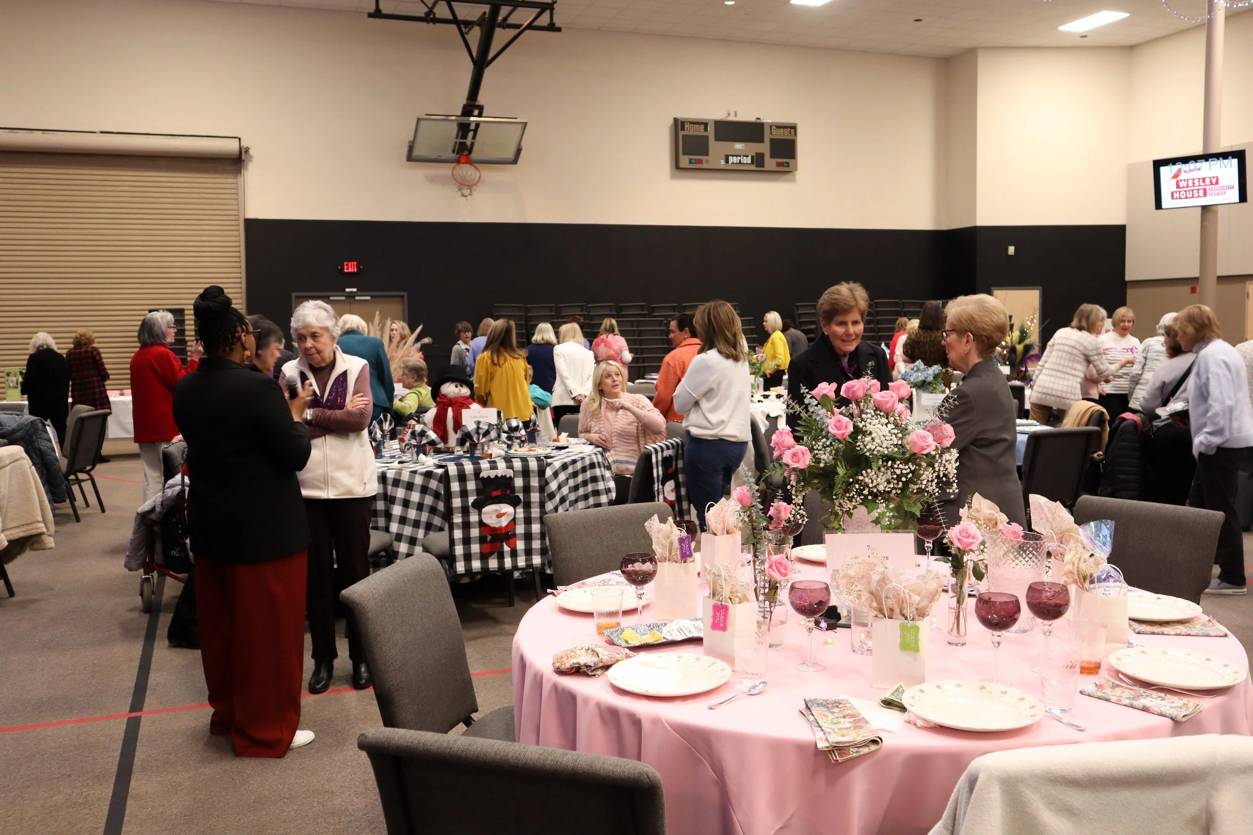 People gathered in a gymnasium during a social event with decorated tables and flower arrangements.