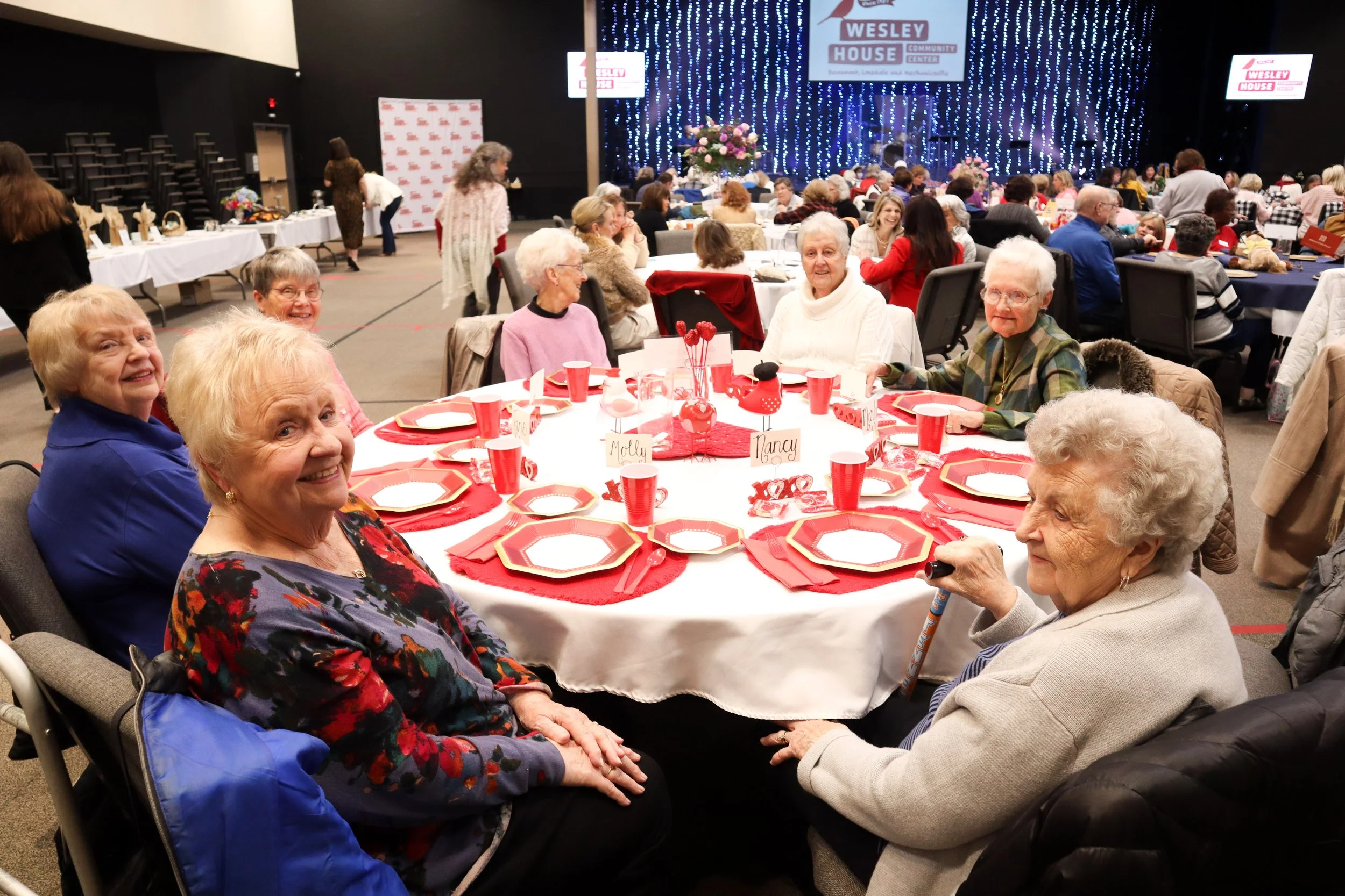 Elderly women sitting around a decorated round table with red and gold plates, cups, and napkins at a community event in a large hall with a stage and blue string lights.