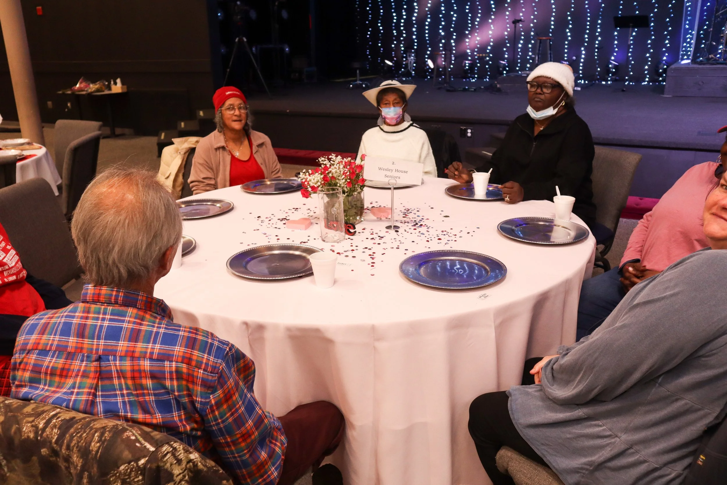 People sitting around a decorated banquet table with a floral centerpiece, plates, cups, and confetti, in front of a stage with string lights; some wear Santa hats and masks, indicating a holiday celebration.