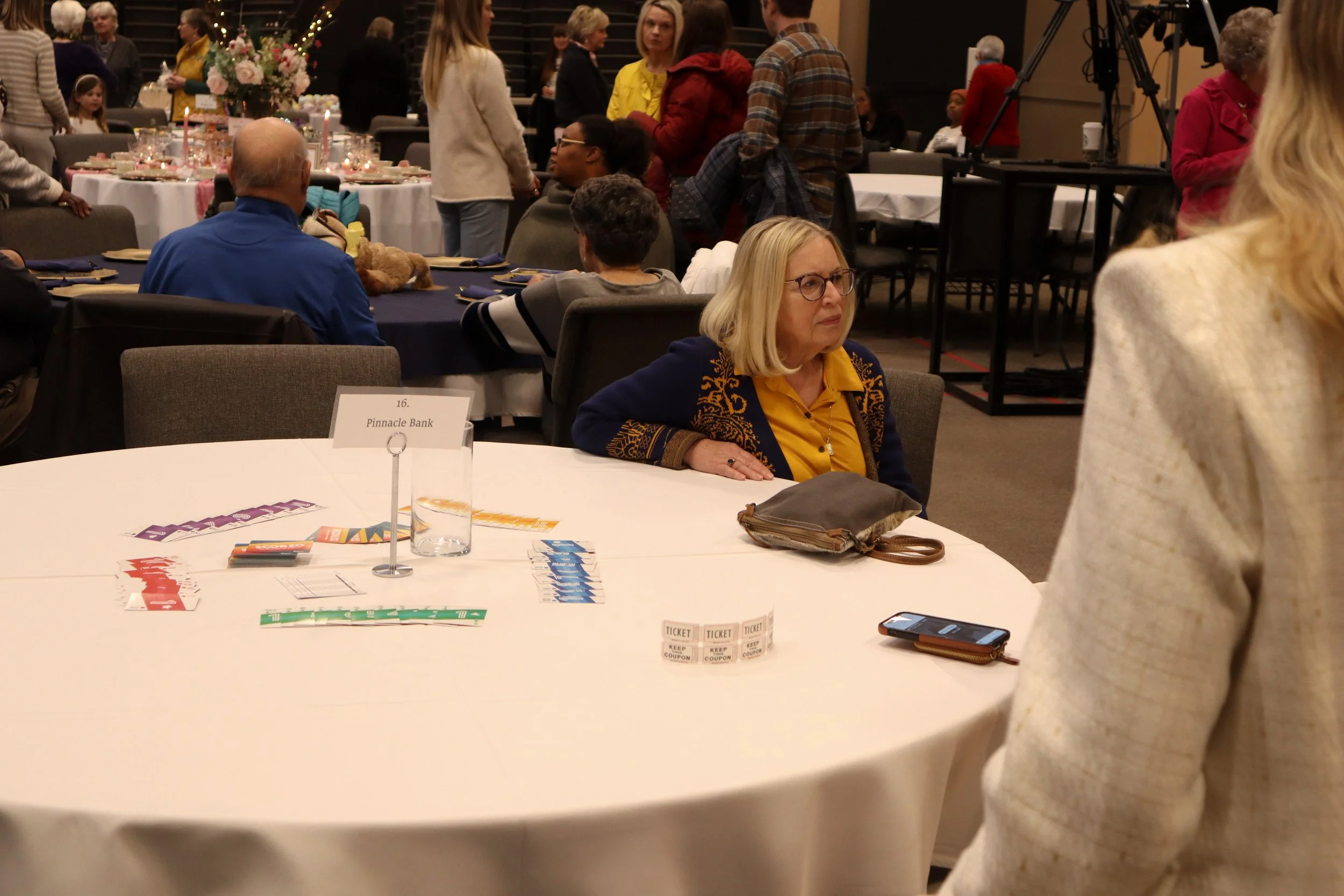 A woman with blonde hair and glasses sitting at a round table during an event, with several people standing and walking around in the background.