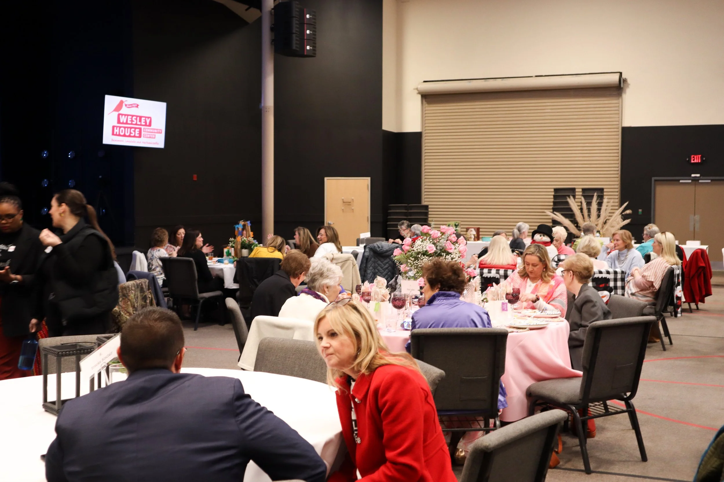 Indoor event with multiple round tables decorated with pink and white flowers, with groups of women sitting and socializing. There is a stage with a digital screen displaying the logo for 'Wesley House'. The room has a black and beige color scheme, w