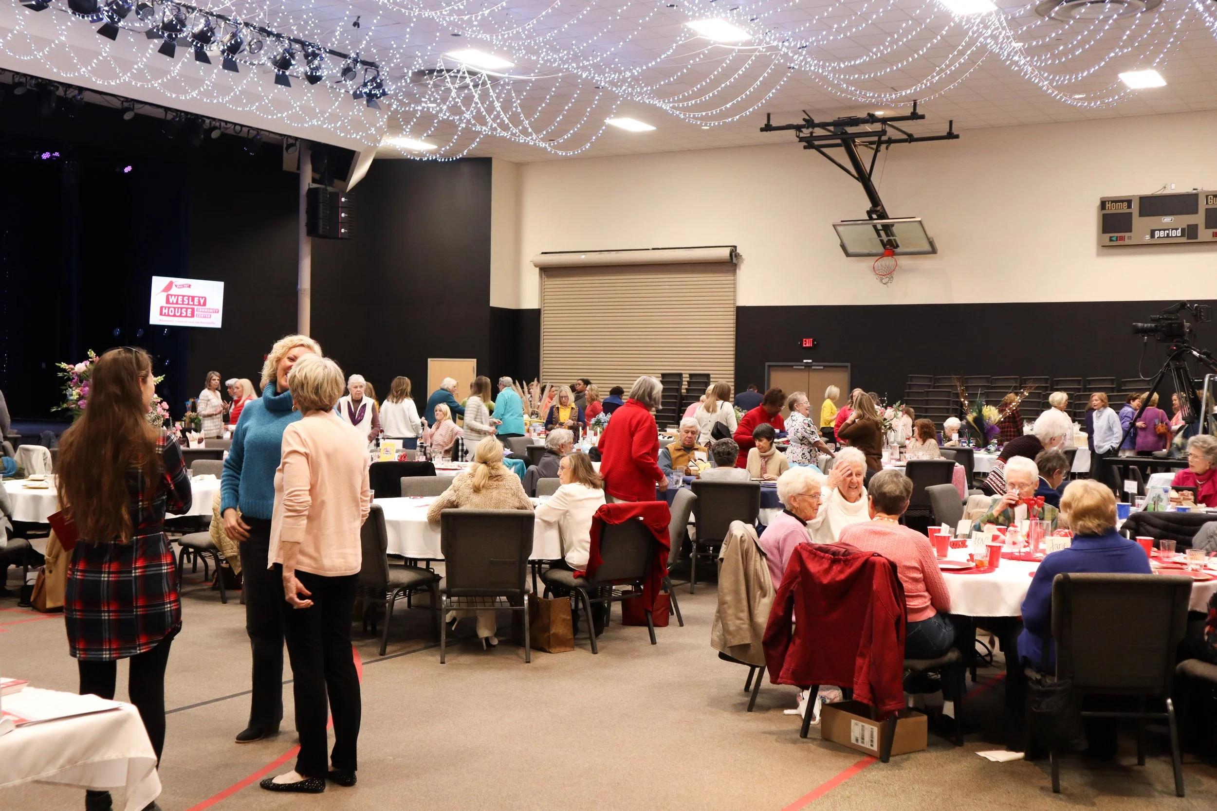 A large indoor event space filled with round tables and chairs, where many elderly people are seated and talking. Some individuals are standing and conversing. The ceiling is decorated with string lights, and a stage is visible in the background with