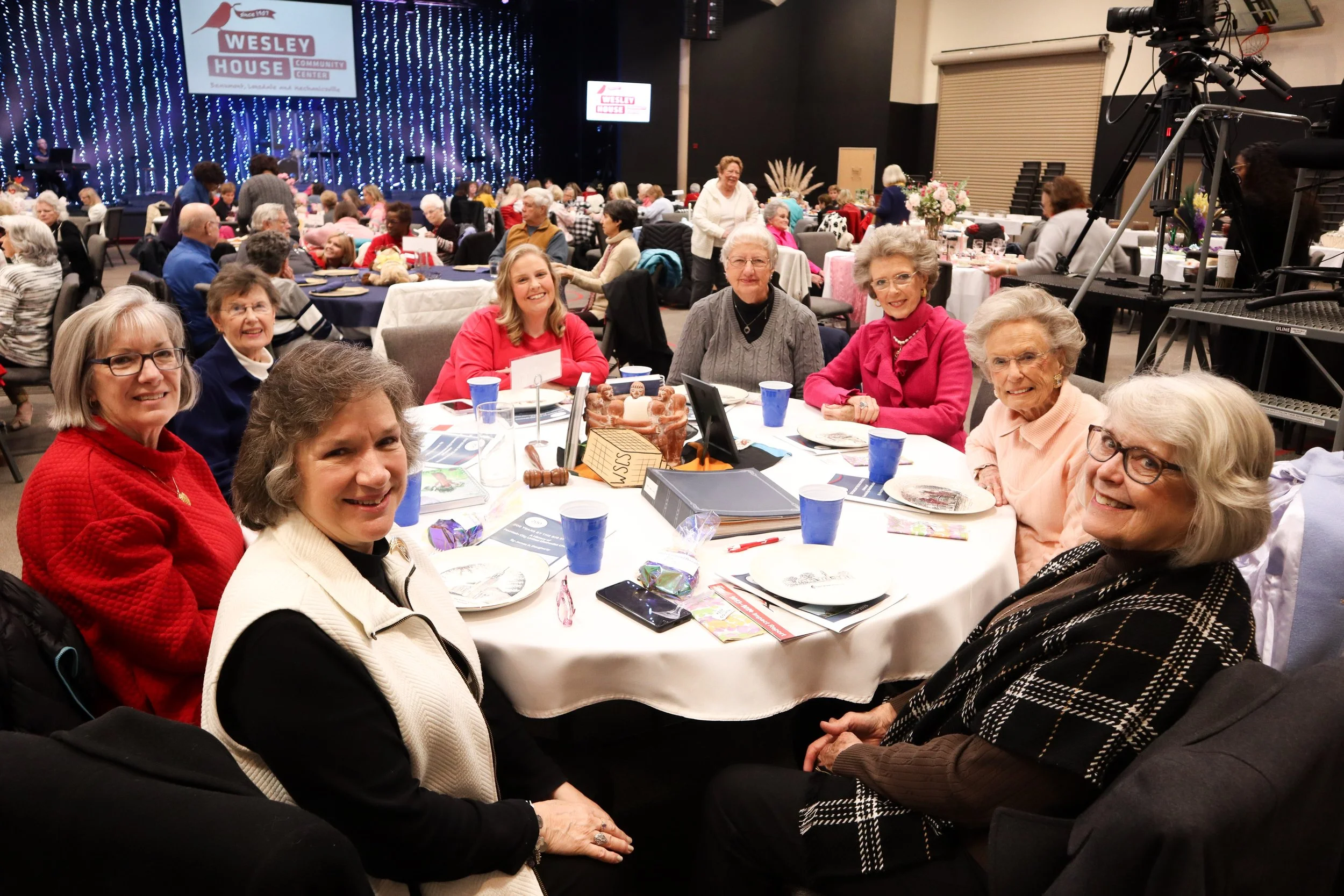 Group of women sitting around a table at a community event in a large hall with a sign that reads 'Wesley House' in the background. The women are smiling and the table is set with plates, cups, and Christmas decorations.