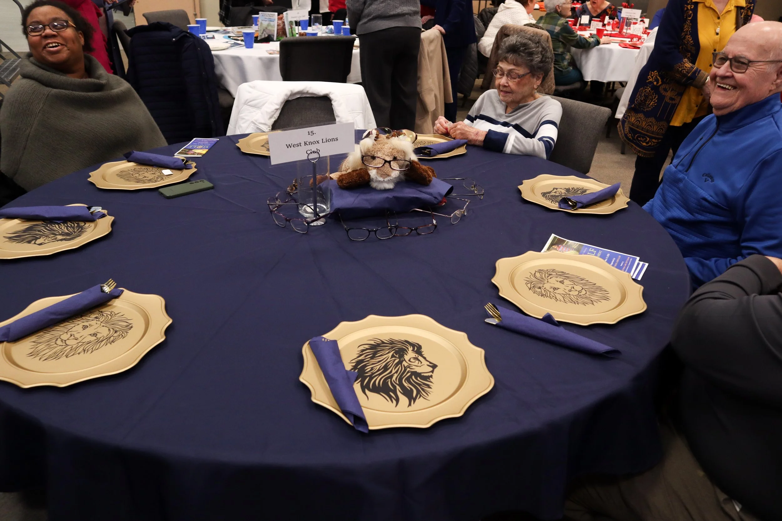 A round table with a navy blue tablecloth decorated with plates featuring a lion's face design, blue napkins, and glasses. In the center, there is a lion-themed plush decoration with glasses on it. Several people are seated around the table, smiling 
