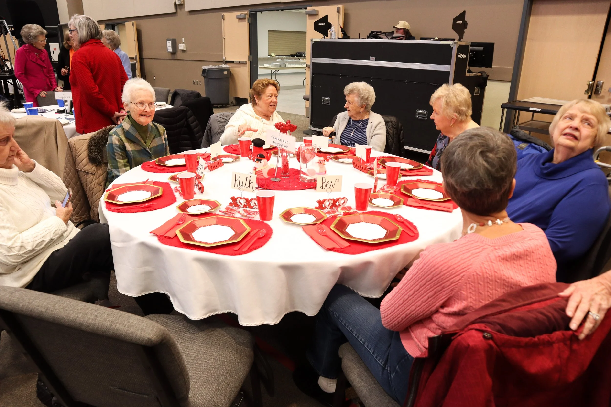 A group of elderly women sitting around a round table decorated for a festive occasion with red and white tableware, holiday-themed decorations, and place cards with names. Some women are talking, one is looking at her phone, and they seem to be in a