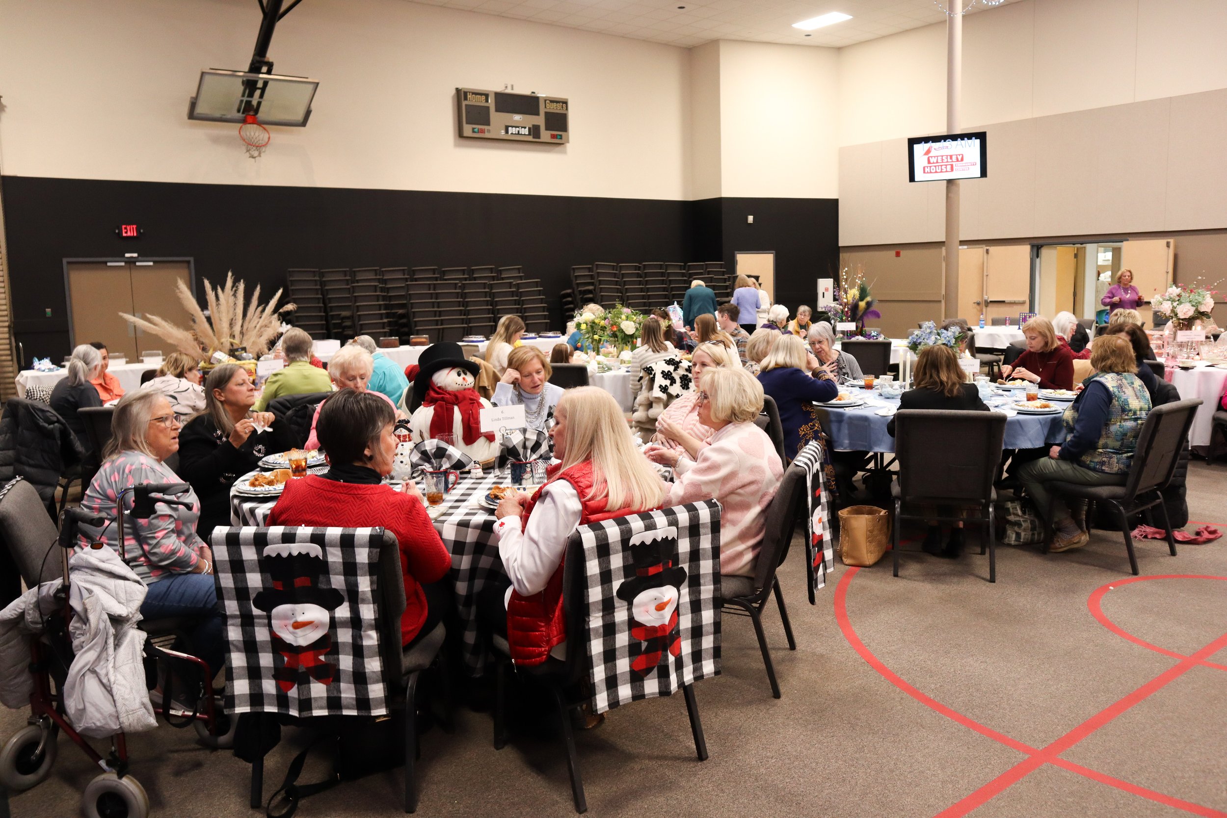 A large indoor gathering with multiple round tables, mostly occupied by women, decorated with winter or holiday-themed napkins and centerpieces, with some tables having Santa or snowman decorations, and a gymnasium or community center setting in the 