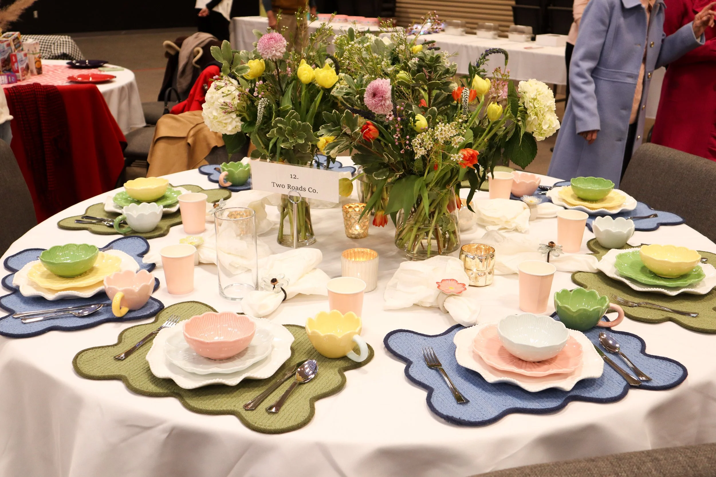 A round banquet table set for a meal with colorful ceramic dishware, including pink, yellow, green, and white bowls and plates, and pink cups. The table has a large centerpiece of mixed flowers in a vase and is decorated with candles and napkins. Sev