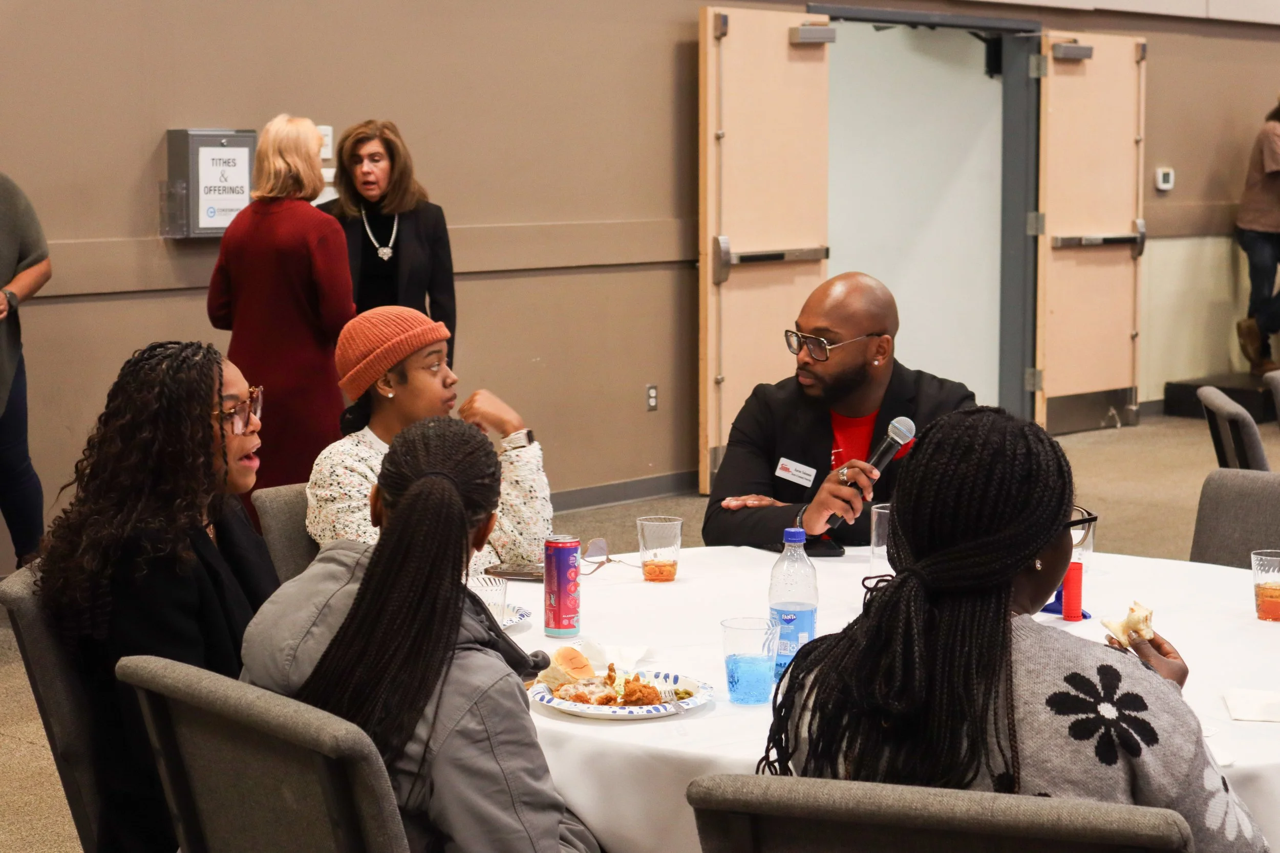 A woman with glasses and braided hair is sitting at a round table with four other women, holding a piece of food, and engaging in a conversation with a man holding a microphone. The table has food, drinks, and a water bottle. Two women are standing i