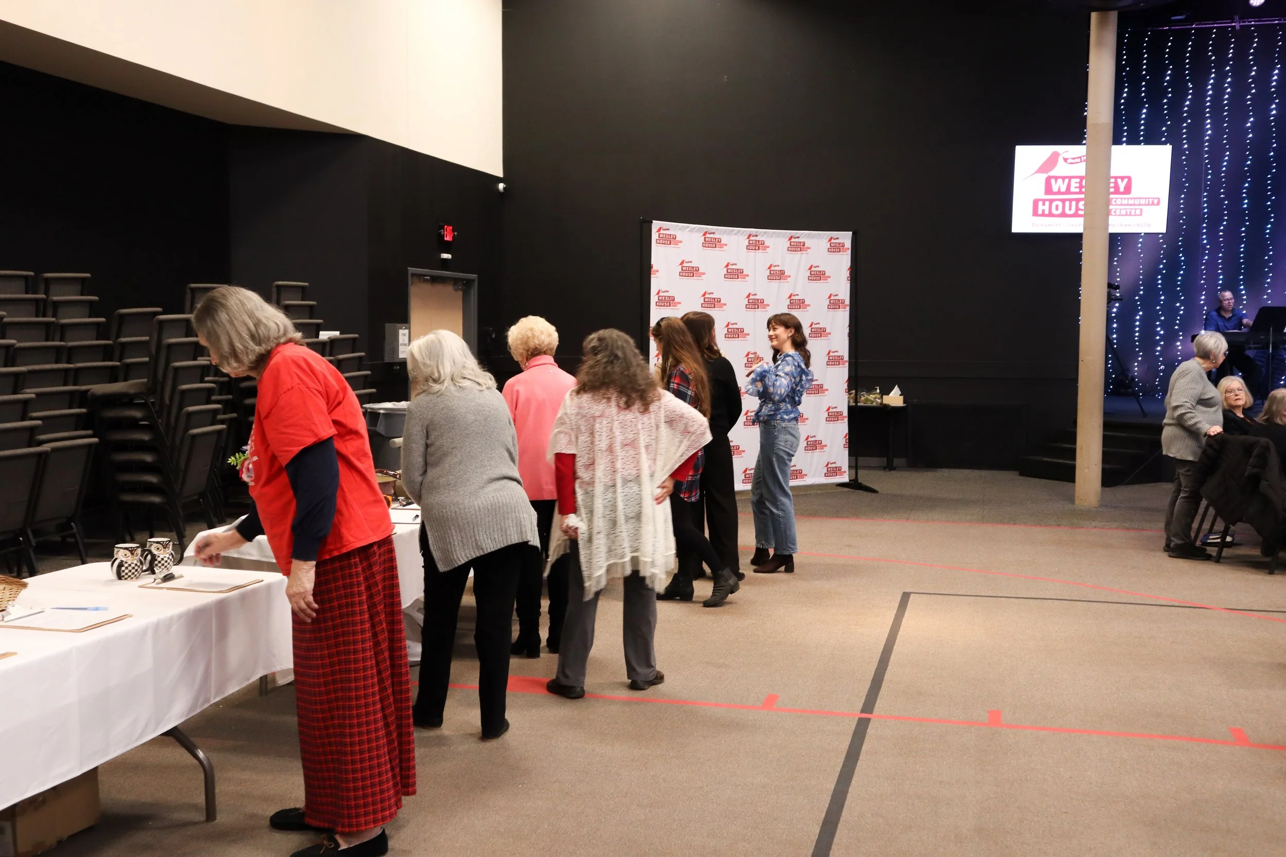 A group of elderly women standing in line at a buffet table in a large event space with a black wall, a stage with a musician, and a screen displaying 'Wesley House Community Center'.