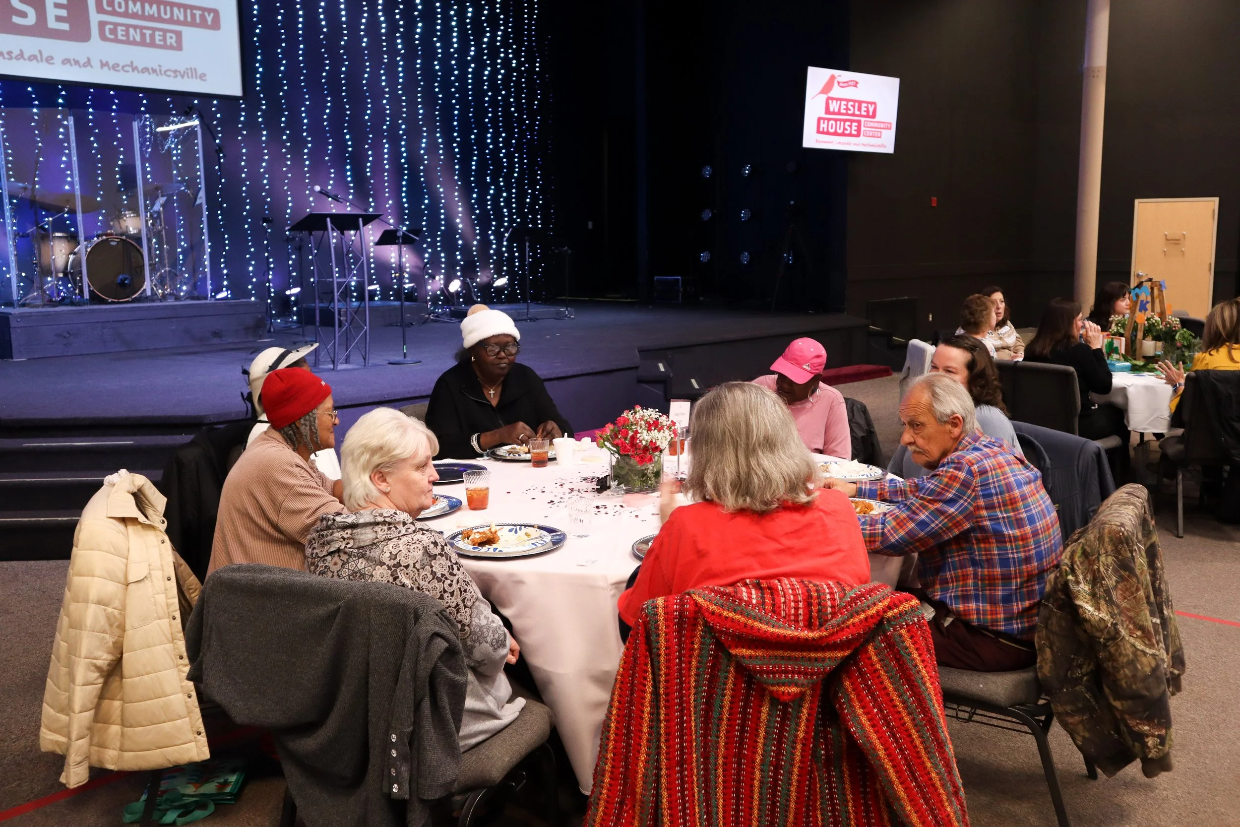 People sitting around a table at a community event, with a stage and holiday decorations in the background.