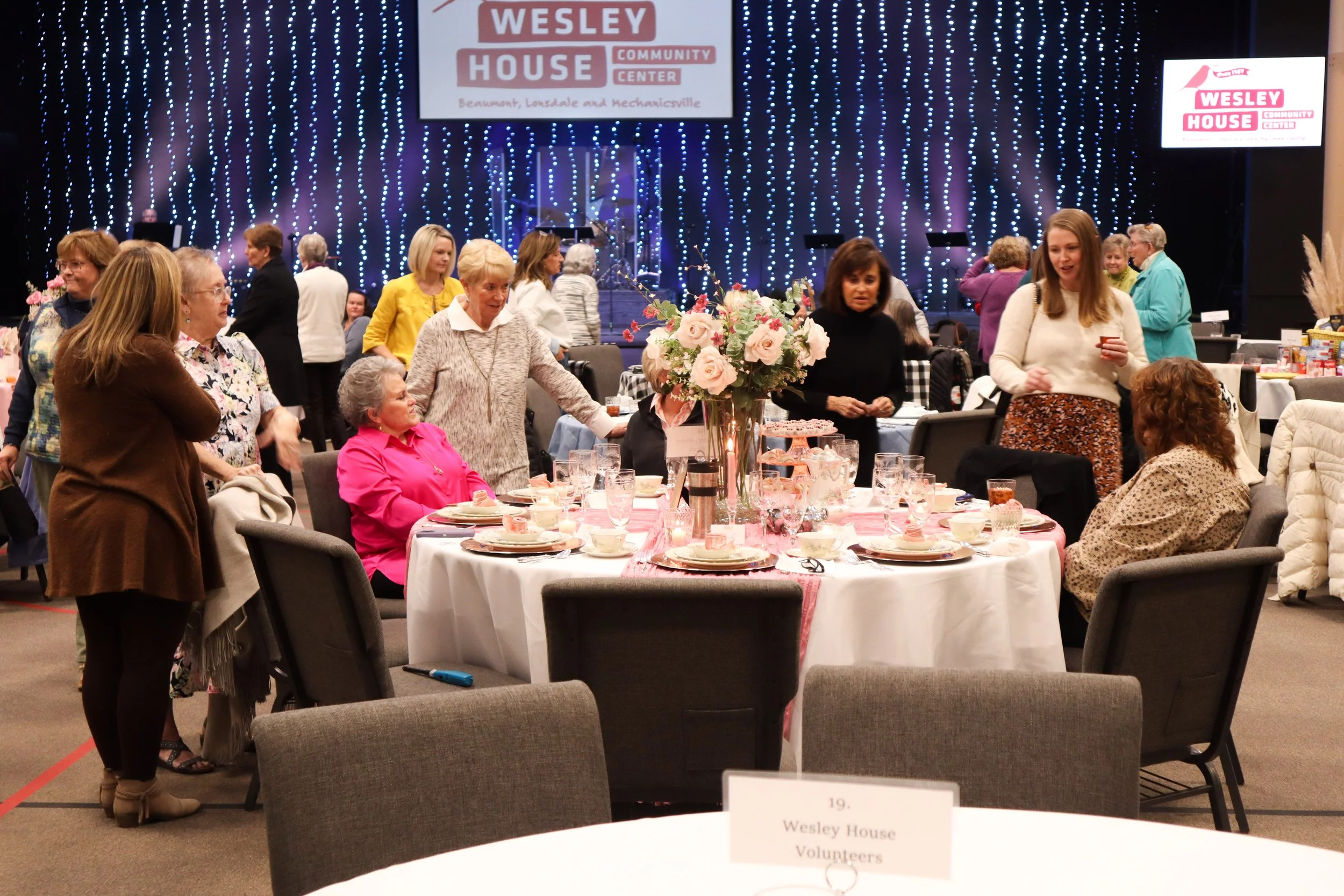 A social gathering at Wesley House Community Center with a round table set with floral centerpiece, teacups, and glasses, surrounded by women engaging in conversation, with a decorated stage in the background featuring hanging string lights and scree