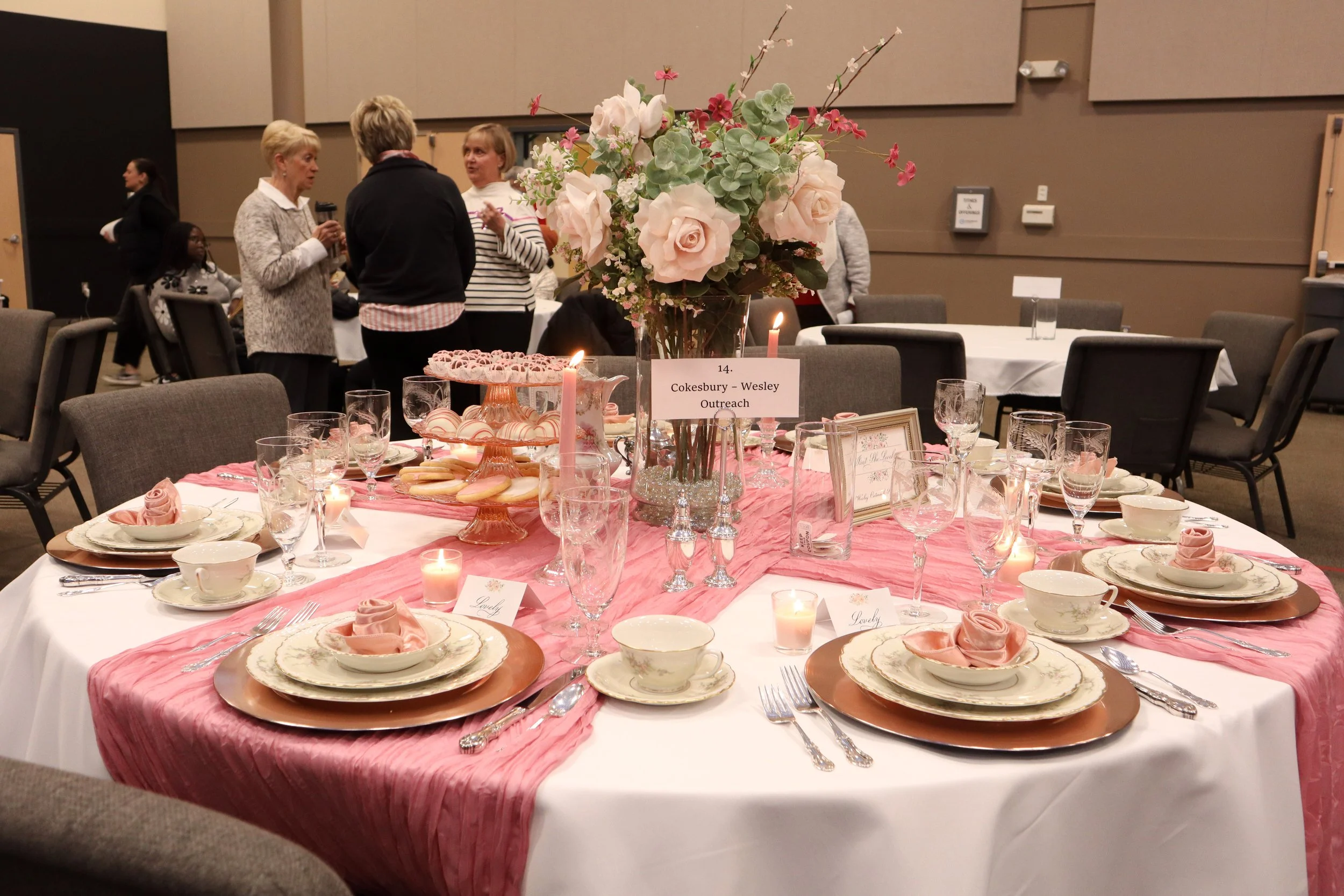 A round banquet table decorated with pink and white floral centerpiece, pink napkins, and elegant tableware, set for a formal event with gift and place cards. In the background, people are standing and talking in a large conference room.