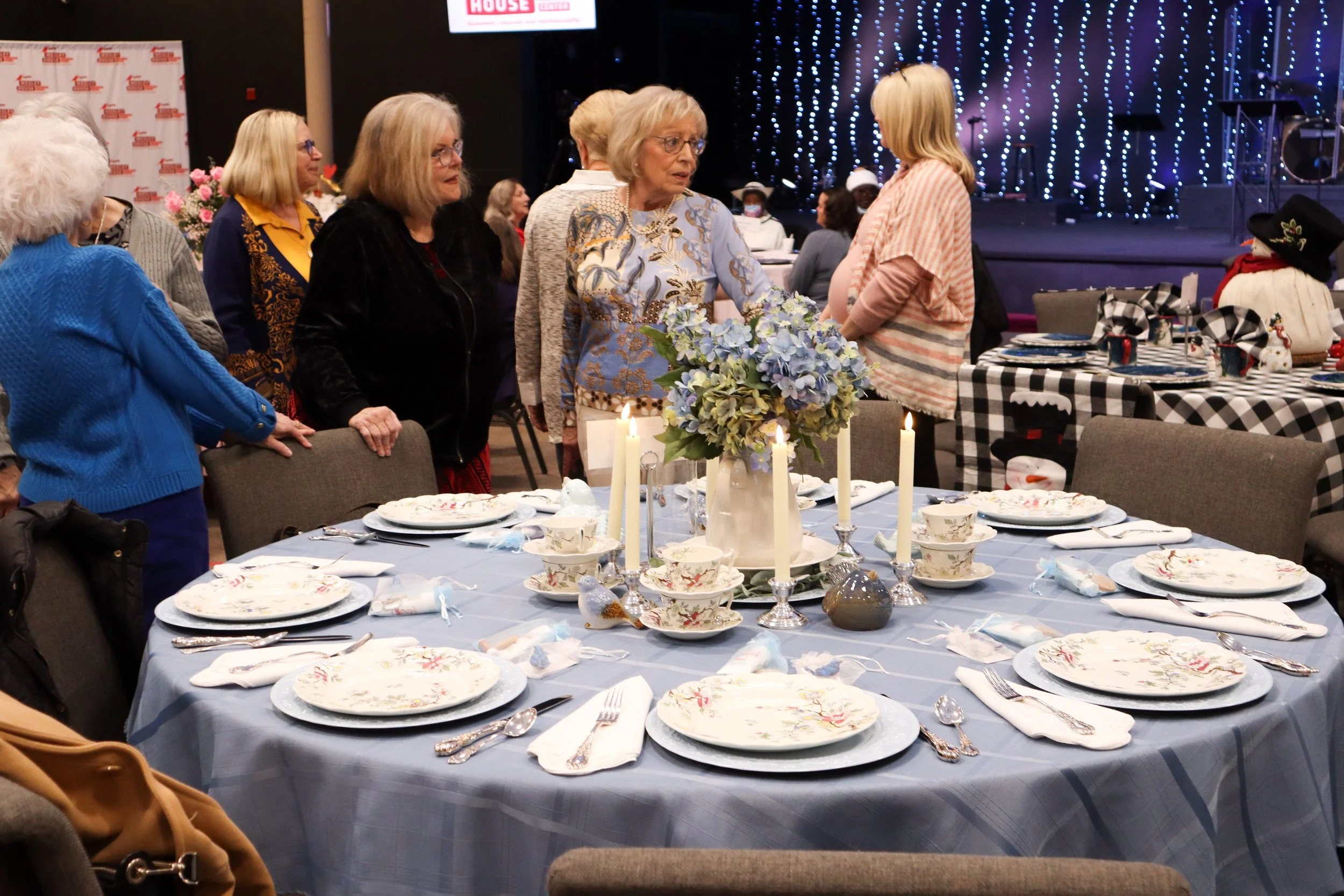 A decorated round table with floral dinnerware, candles, and a floral centerpiece in a banquet hall. Several women are standing and conversing near the table, with more tables and a stage decorated with blue lights in the background.