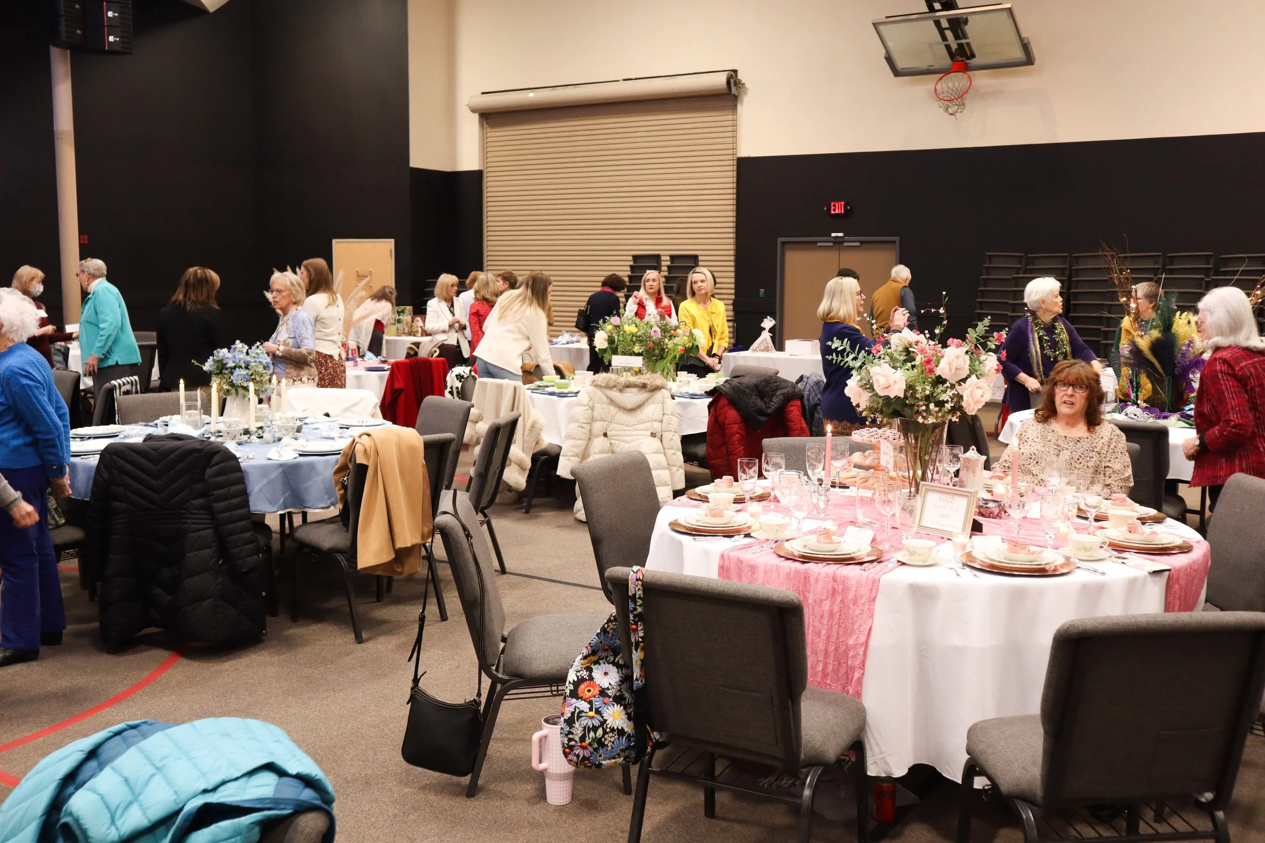 A gathering of women in a gymnasium decorated with floral centerpieces and set for a meal, with tables covered in pink and white tablecloths.