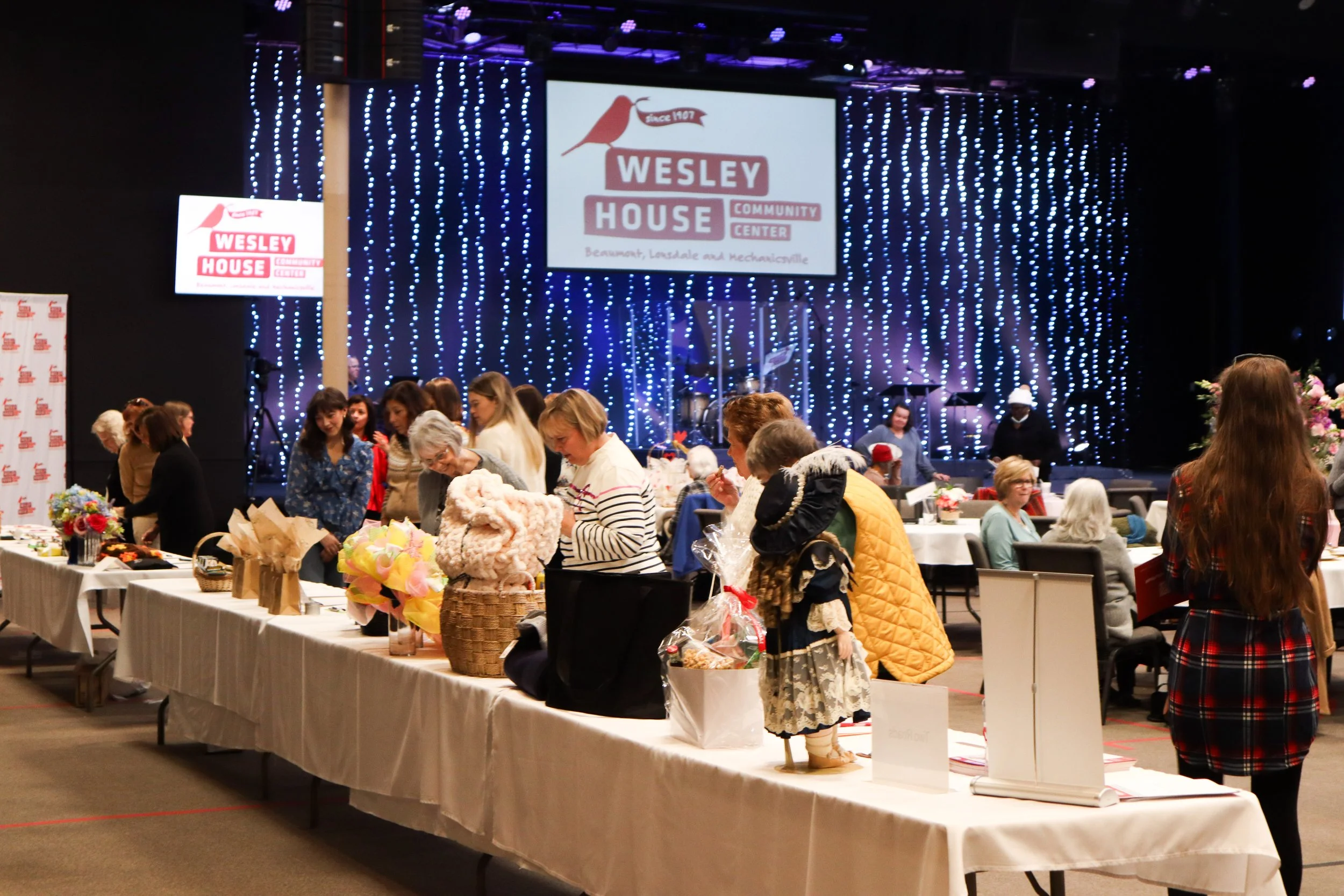 People gathered around tables at a community event in a large hall with a decorated stage in the background, displaying the Wesley House Community Center logo.