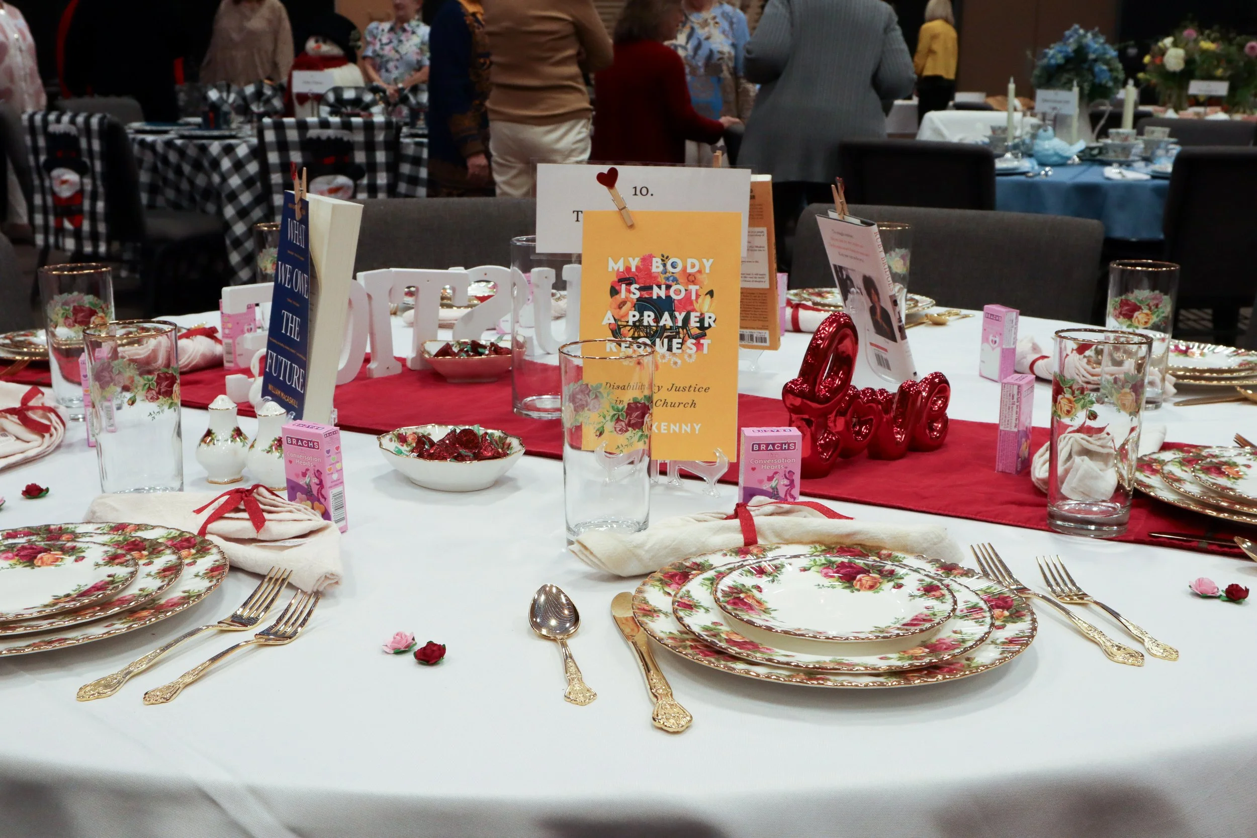 Round banquet table decorated for a celebration with floral patterned plates, gold flatware, white napkins tied with red ribbons, and glassware with floral designs. The table has a red table runner, decorative signs, and a red 