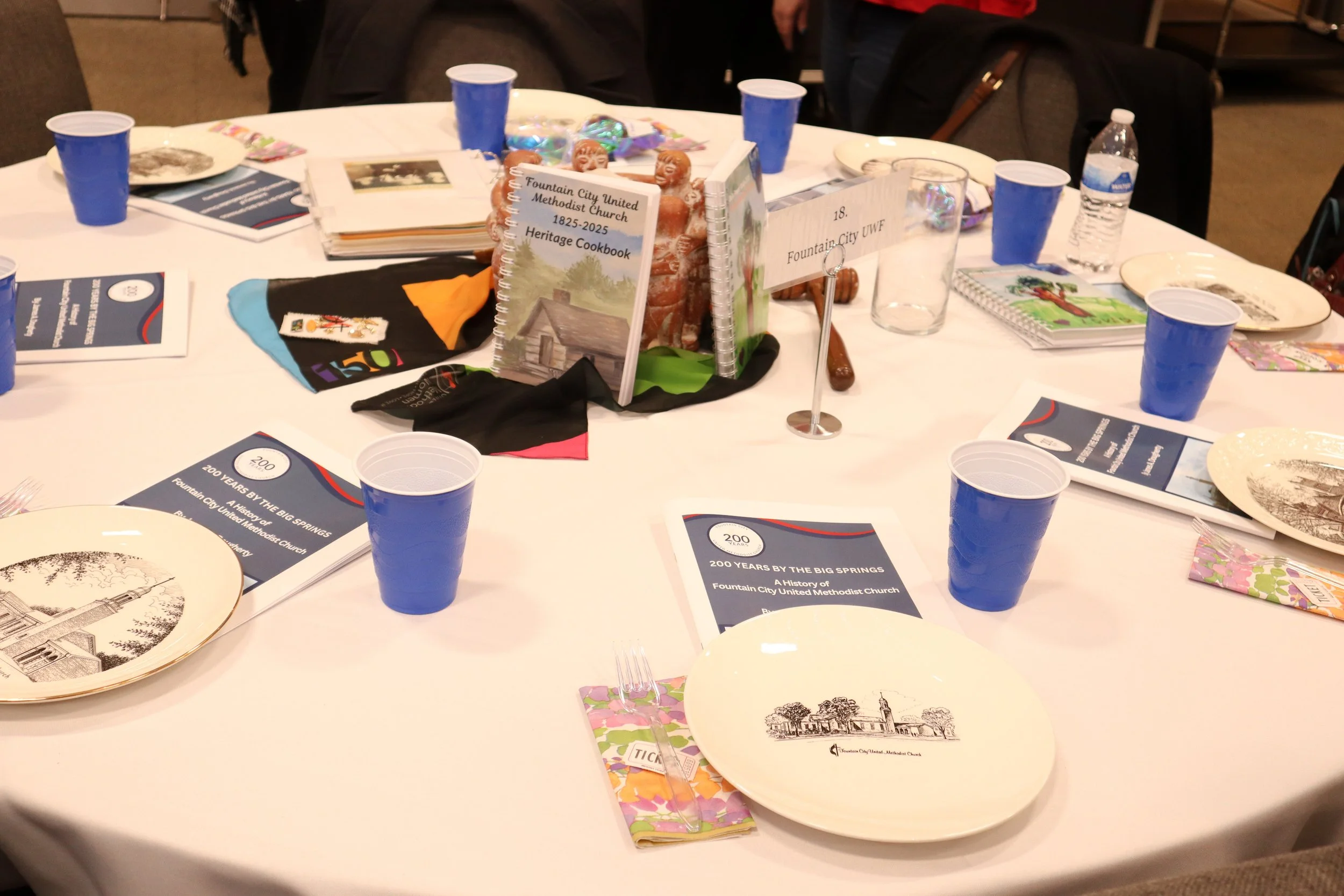 A round table set for a celebration with pamphlets, plates, blue cups, and a centerpiece featuring a cookbook and decorations, celebrating 200 years of Fountain City United Methodist Church.