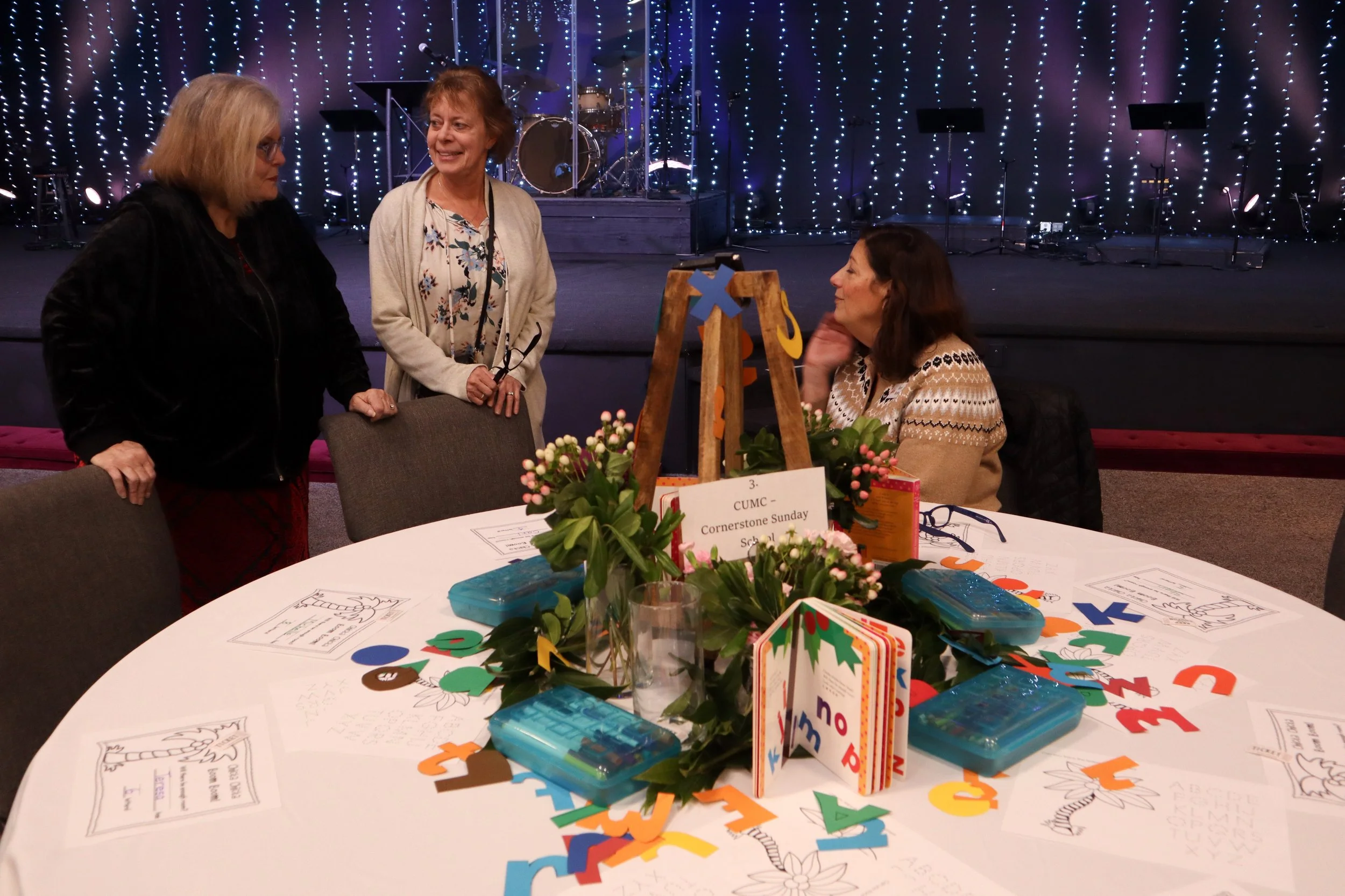 Three women gathered around a table decorated with holiday themed crafts and flowers at a Cornerstone Sunday School event, with a stage and musical instruments in the background.
