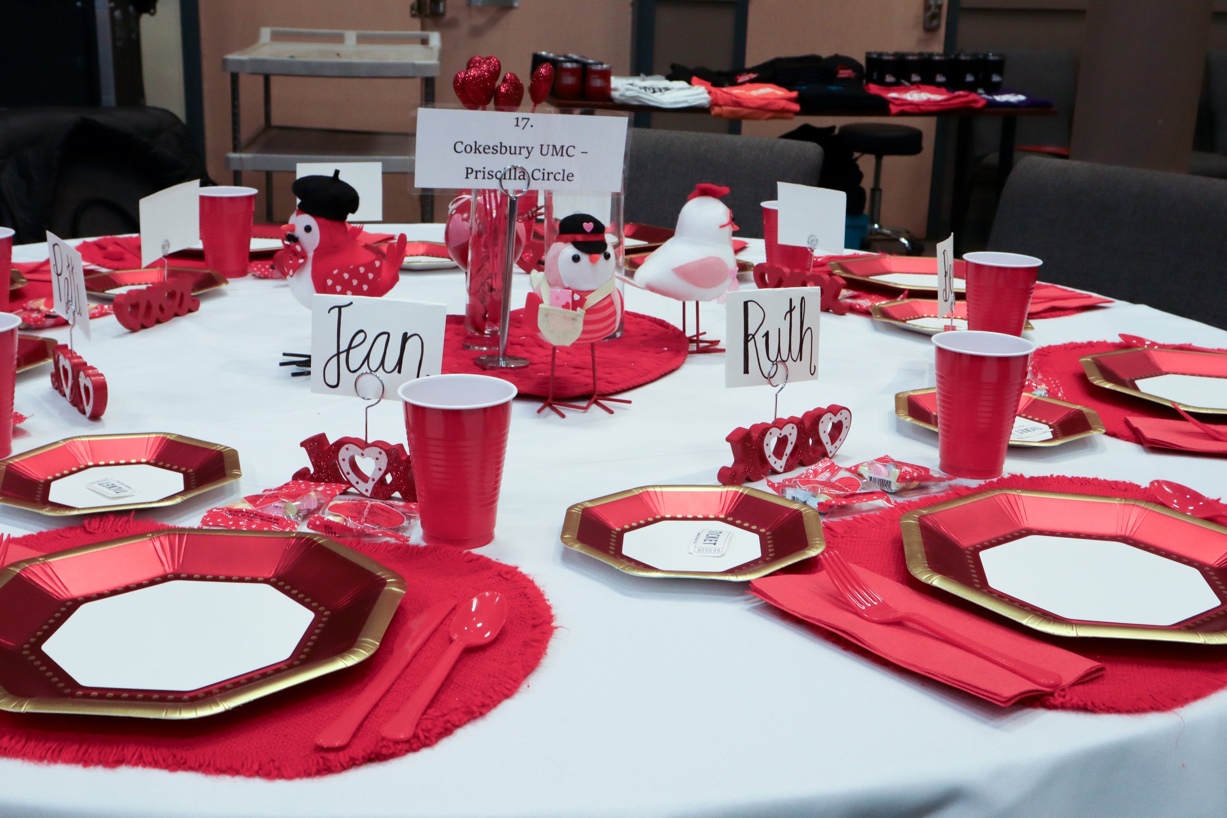Valentine's Day themed table with red and gold plates, red cups, red napkins, and heart-shaped decorations. There are plush chicken and duck centerpieces and handwritten name cards for guests.