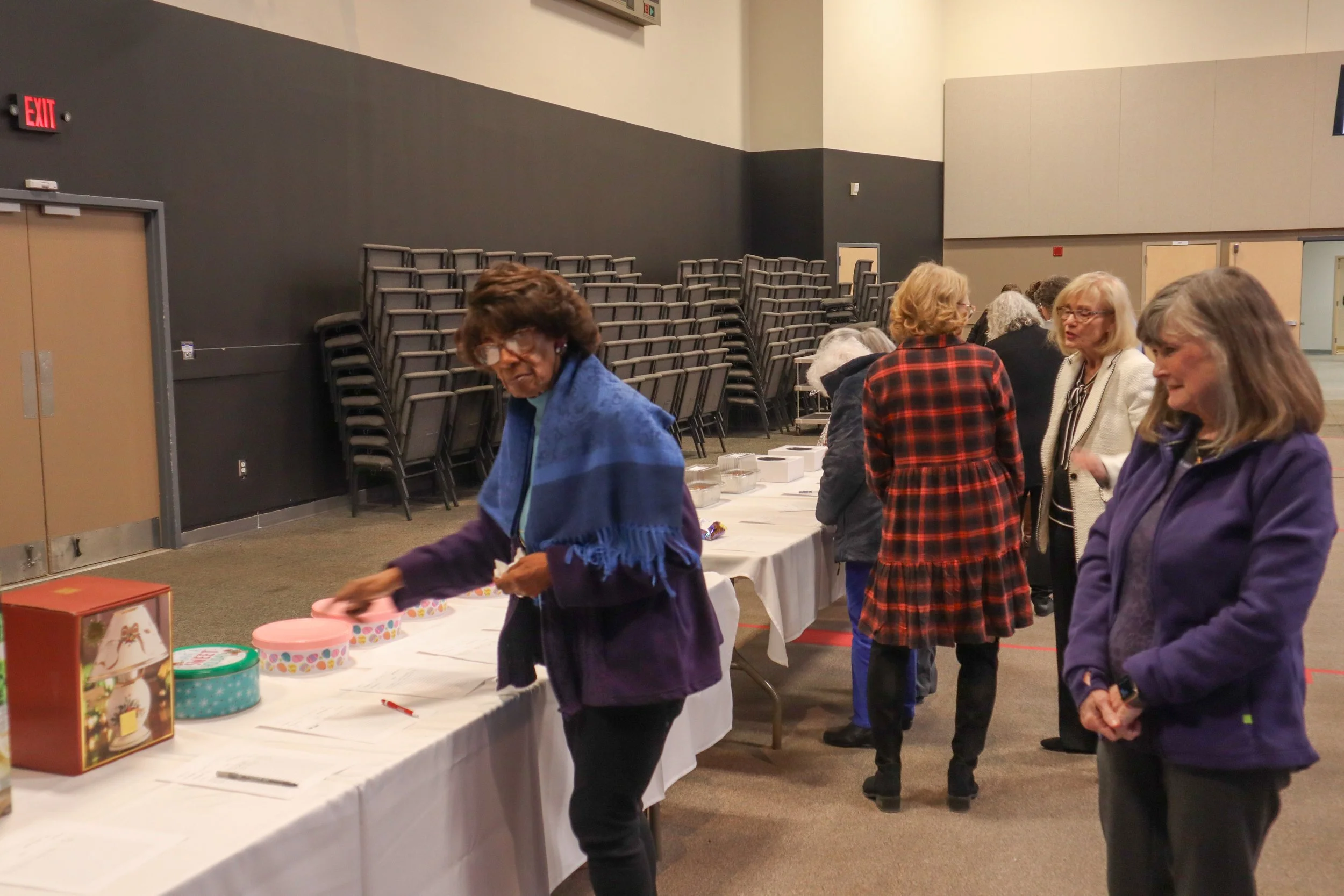 Women gathered in line at a table with baked goods and gift boxes in a large room with stacked chairs and an exit sign.