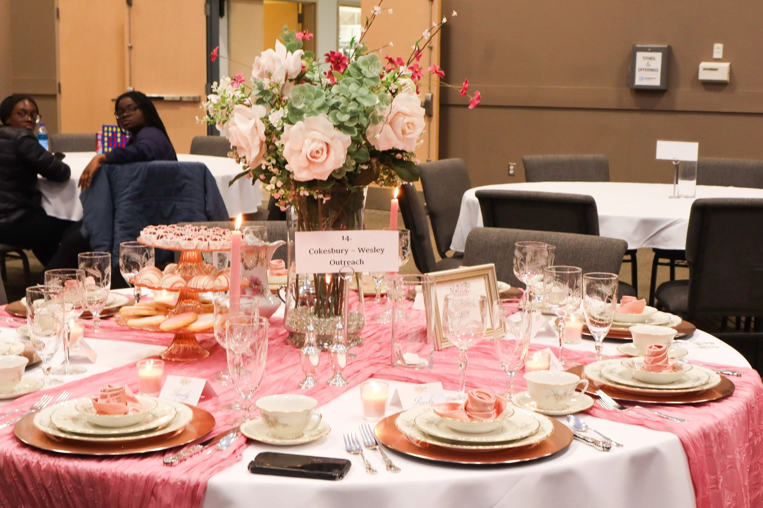 A round banquet table decorated with a pink table runner, fine china, glassware, and pink napkins. The center features a large floral arrangement with pink roses and greenery, and lit pink candles. In the background, there are empty chairs and a few 