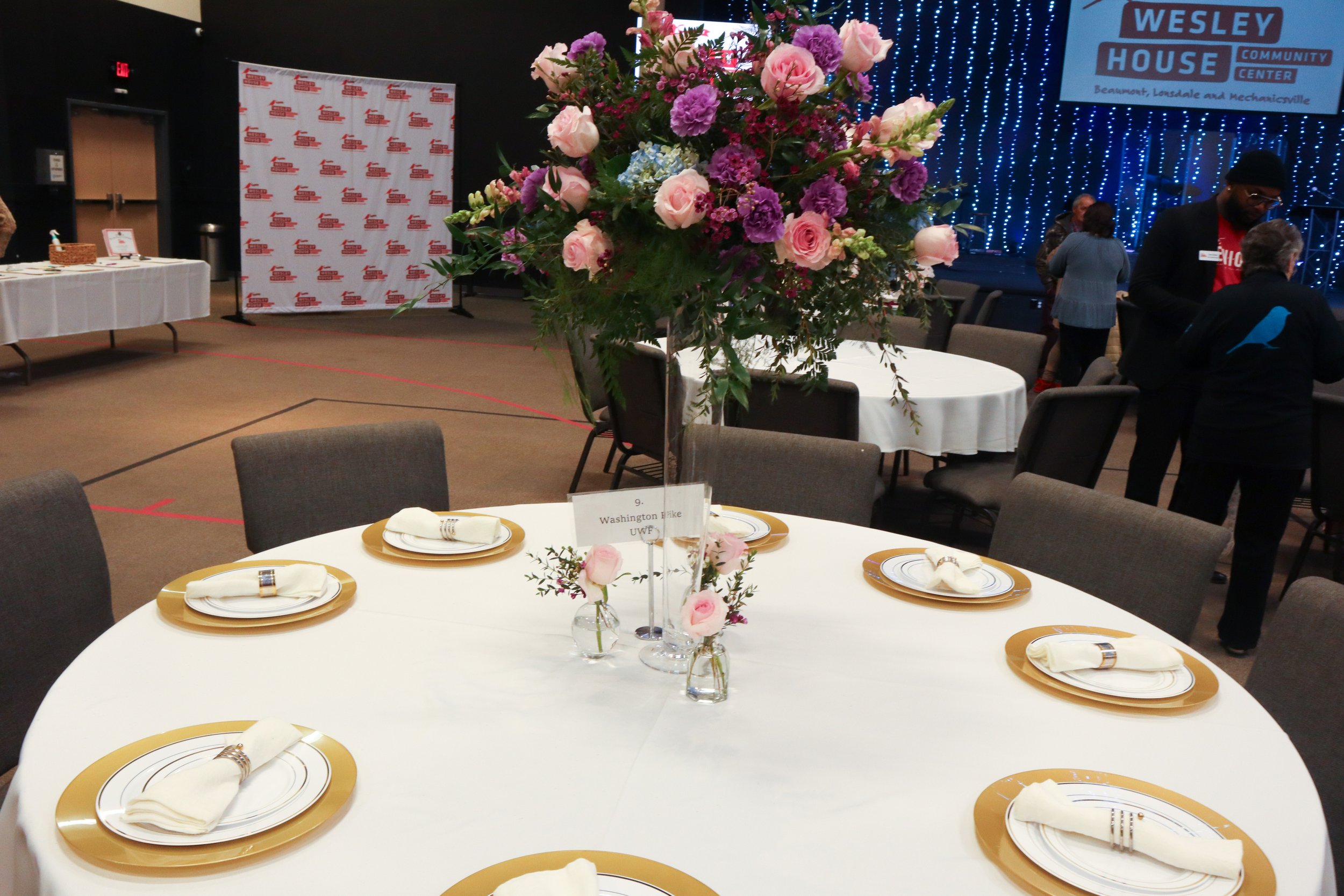 A round table set for an event with gold and white plates, white napkins, and a floral centerpiece with pink and purple flowers in a clear vase. In the background, there are blue string lights, a banner, and a few people standing and talking.