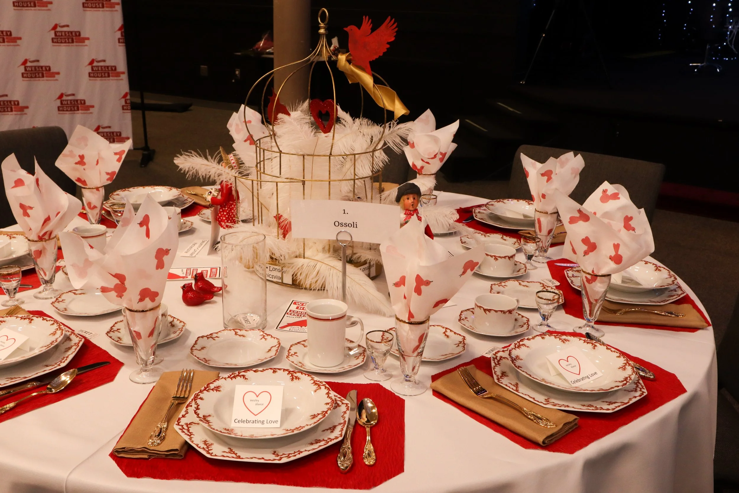 A round table set for a holiday celebration with white and red dinnerware, gold utensils, and folded napkins with red Bunny patterns. The table has a central decorative display with a golden cage, white feathers, red Dove and heart ornaments, and sma