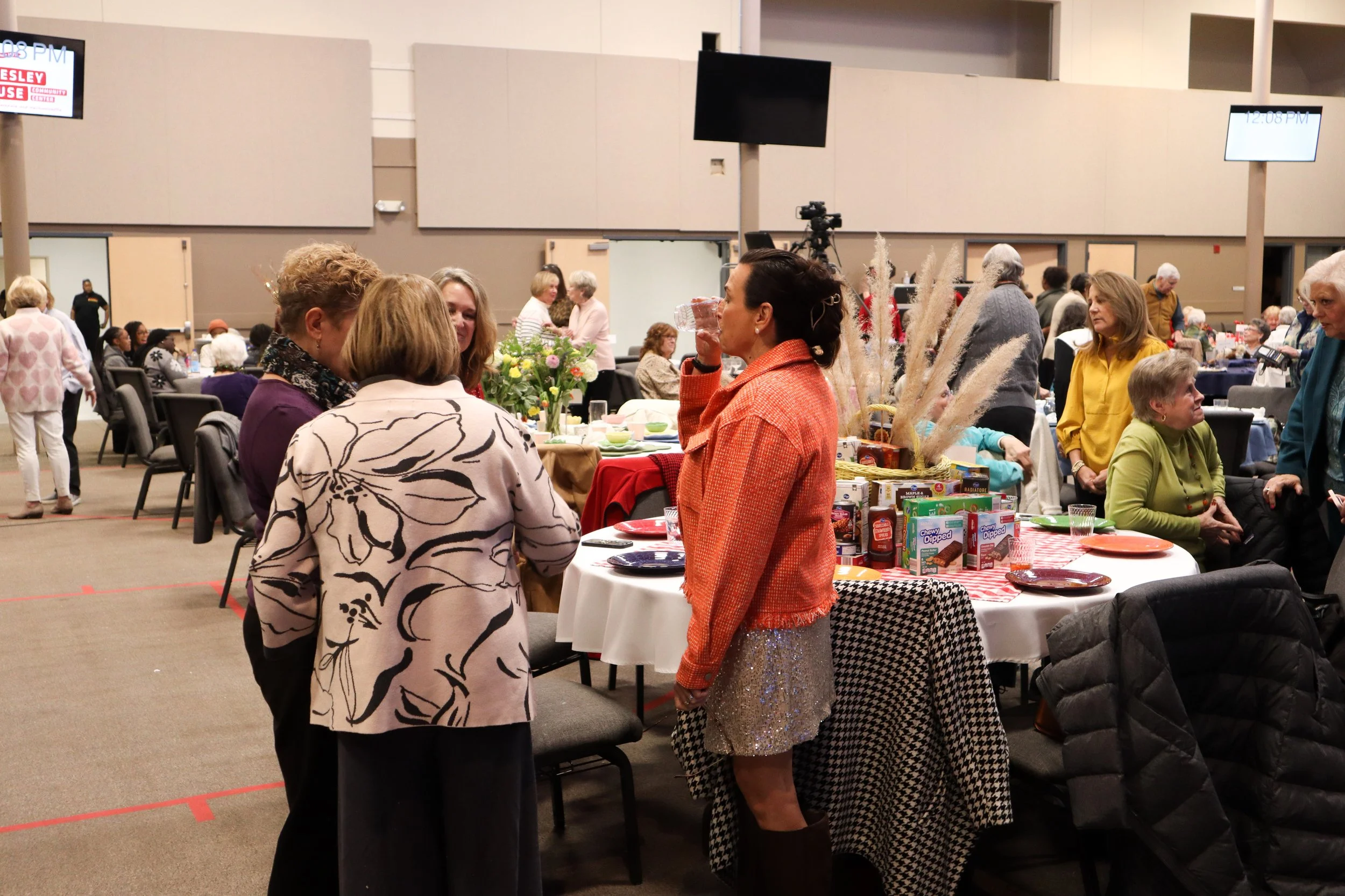 People socializing at a gathering with decorated tables, food, and flowers in a large indoor event space.
