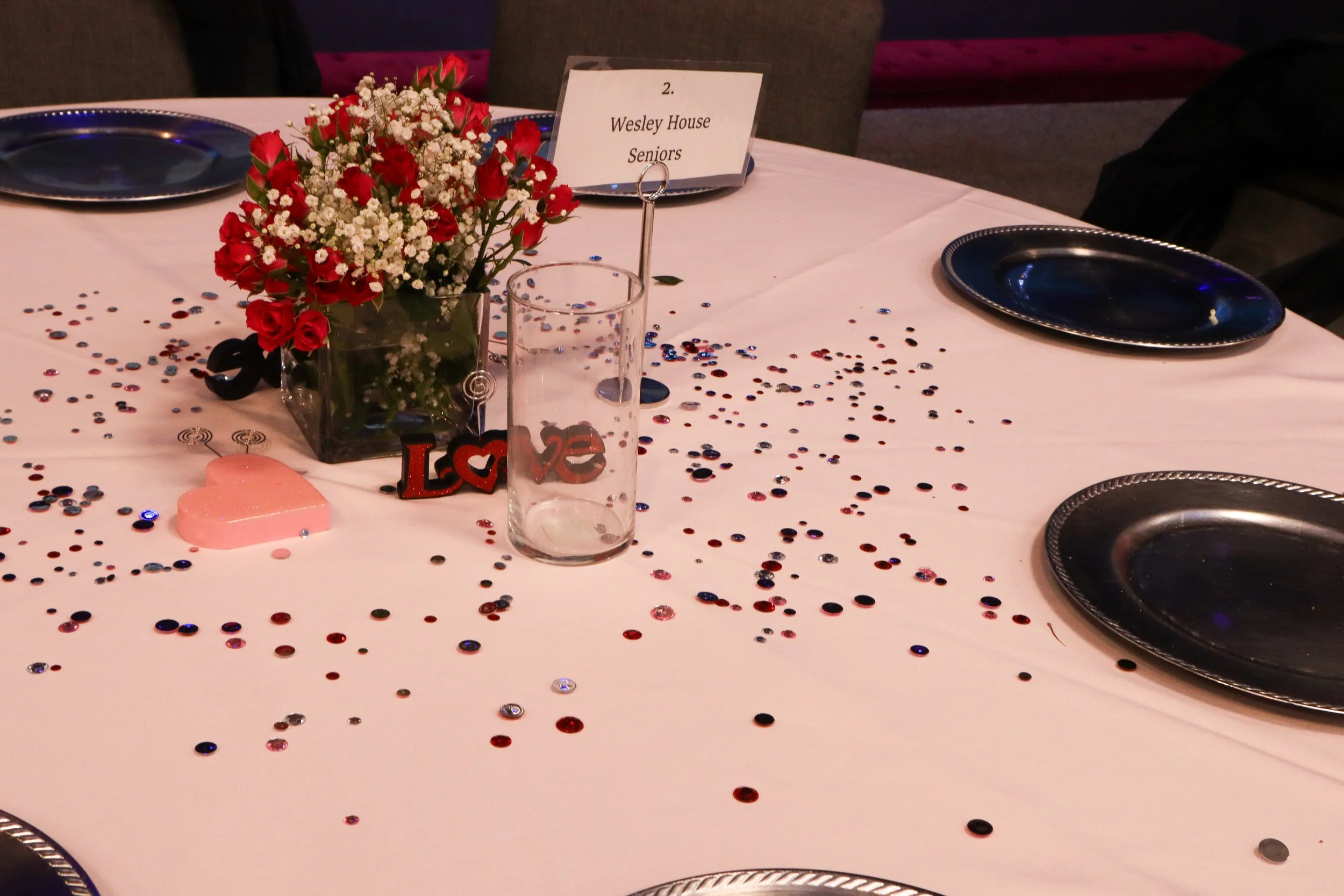 A round banquet table decorated with red, white, and pink flowers in a glass vase, surrounded by scattered red and black confetti, a pink heart-shaped candle, a decorative 'Love' sign, a clear glass, and several dark blue plates. There is a small sig