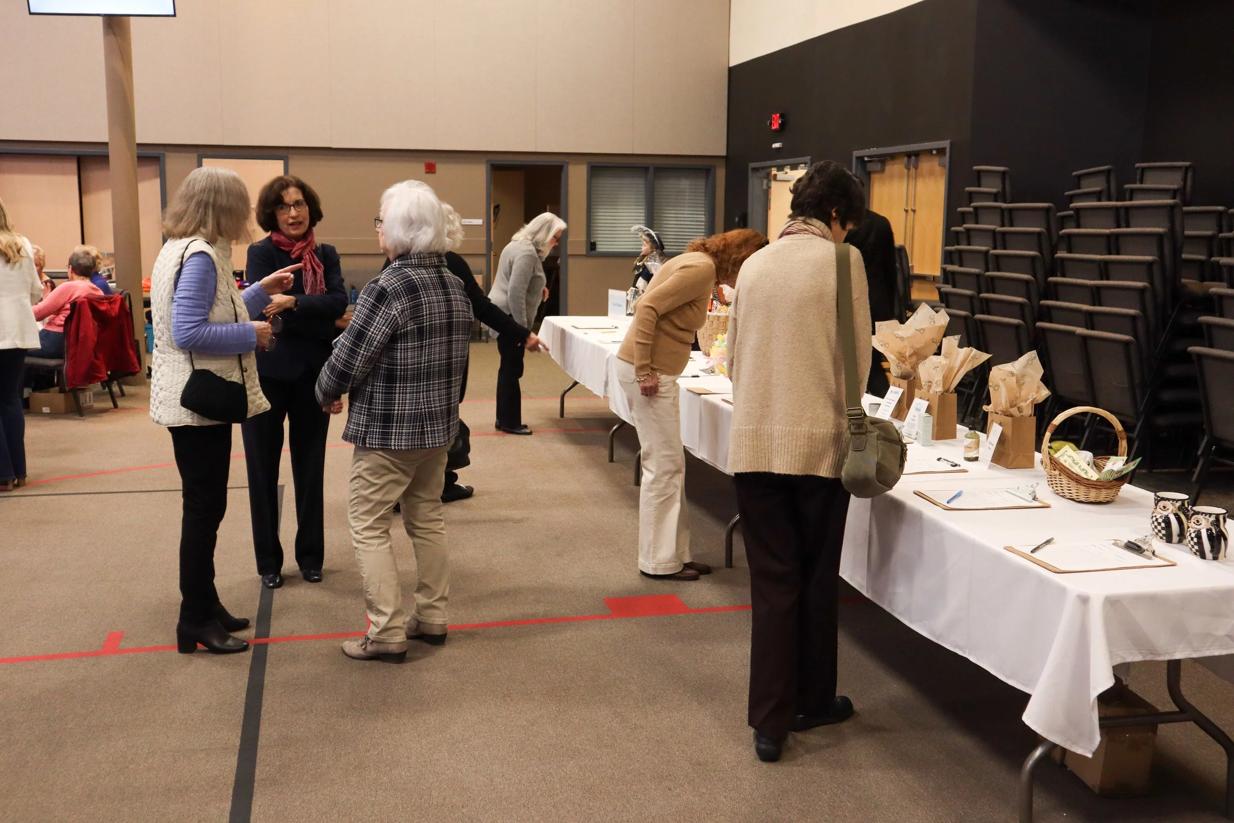 People socializing and shopping at a craft fair or market in an indoor venue with display tables and stacked chairs.
