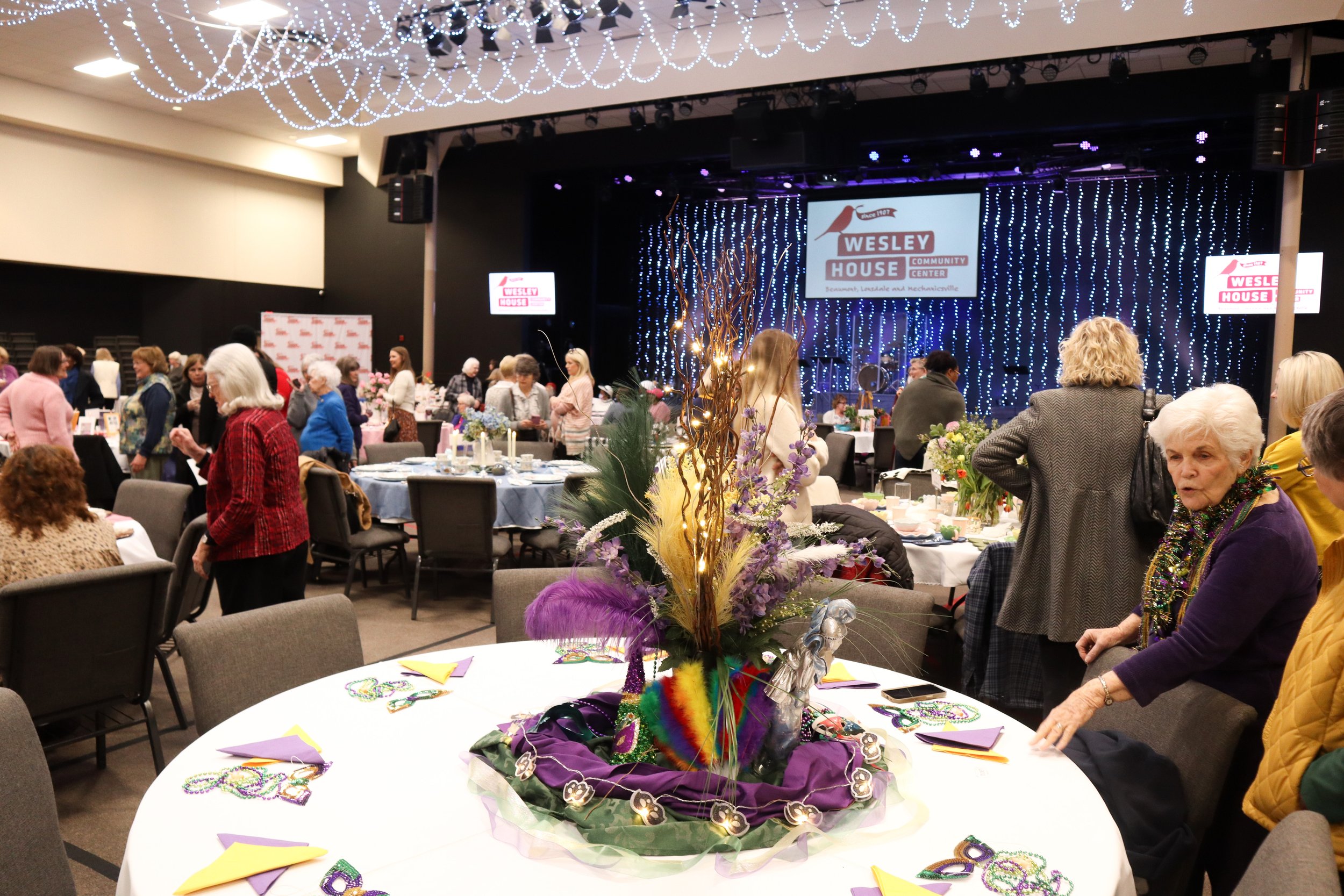 A large indoor event with many women gathered around tables decorated with Mardi Gras beads and colorful napkins. The venue has a stage in the background with a sign that reads 'Wesley House Community Center' and decorative string lights hanging from