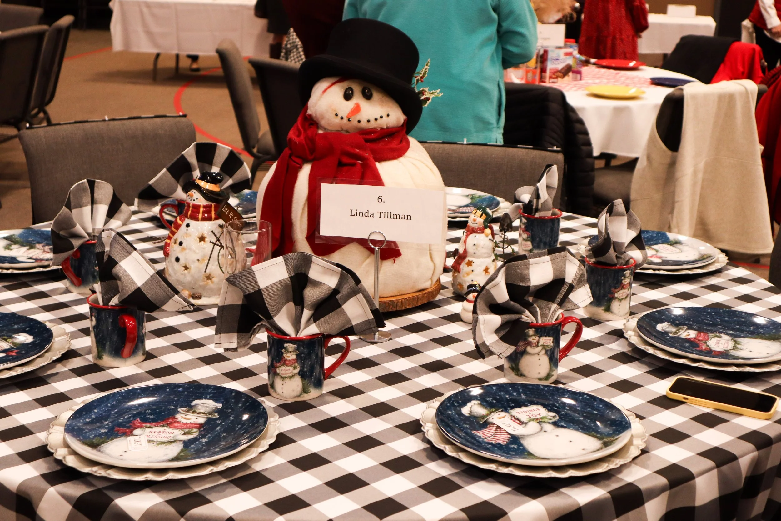 A holiday table setting with a large snowman centerpiece, surrounded by snowman-themed plates and mugs with black-and-white checkered napkins. A sign on the table reads 'Linda Tillman,' and there are other tables and people in the background.