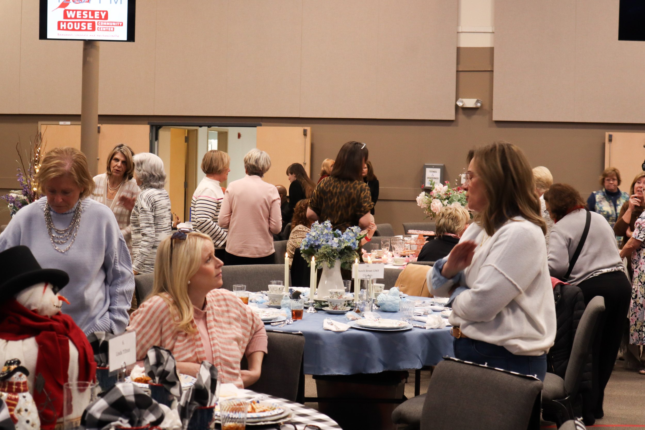 Indoor gathering of women at a decorated table with floral centerpieces, plates, glasses, and holiday decorations, in a community center or banquet hall.
