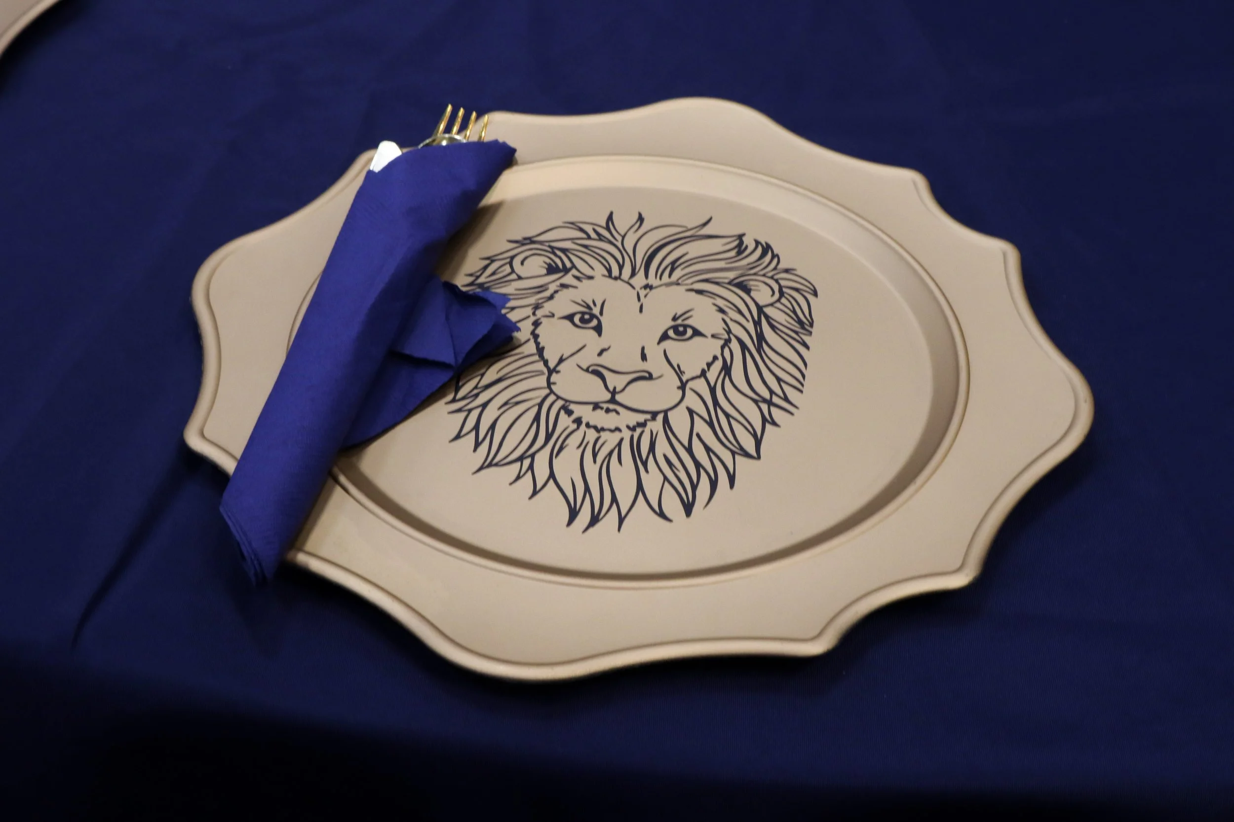 A decorative place setting with a cream-colored plate featuring a black line drawing of a lion's face, placed on a dark blue tablecloth, with a blue napkin and gold-colored cutlery.