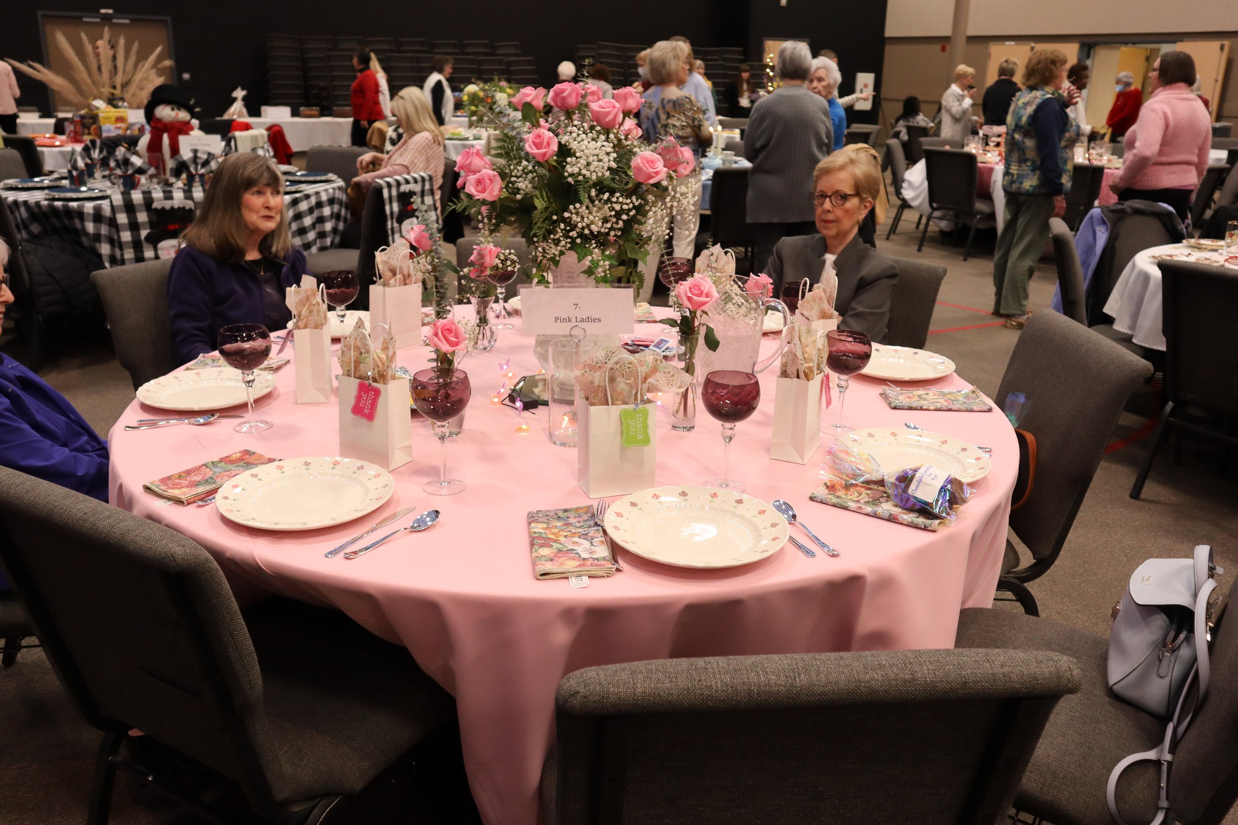 Round table with pink tablecloth and floral centerpiece at a social event, surrounded by people, with place settings, glasses, gifts, and napkins.