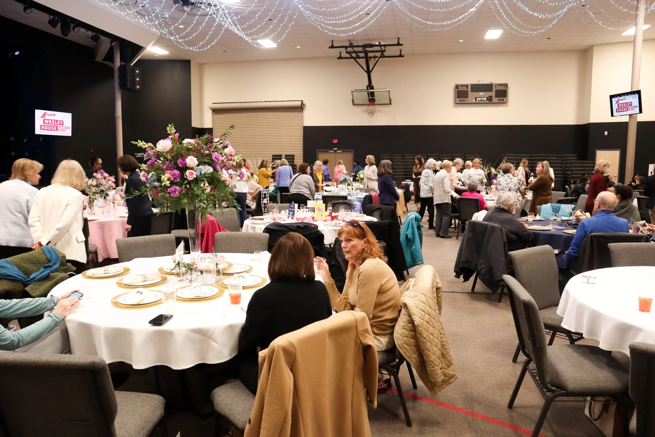 People gathered in a large hall decorated for an event, with round tables set with plates, glasses, and floral centerpieces, and some people sitting and talking while others are mingling.