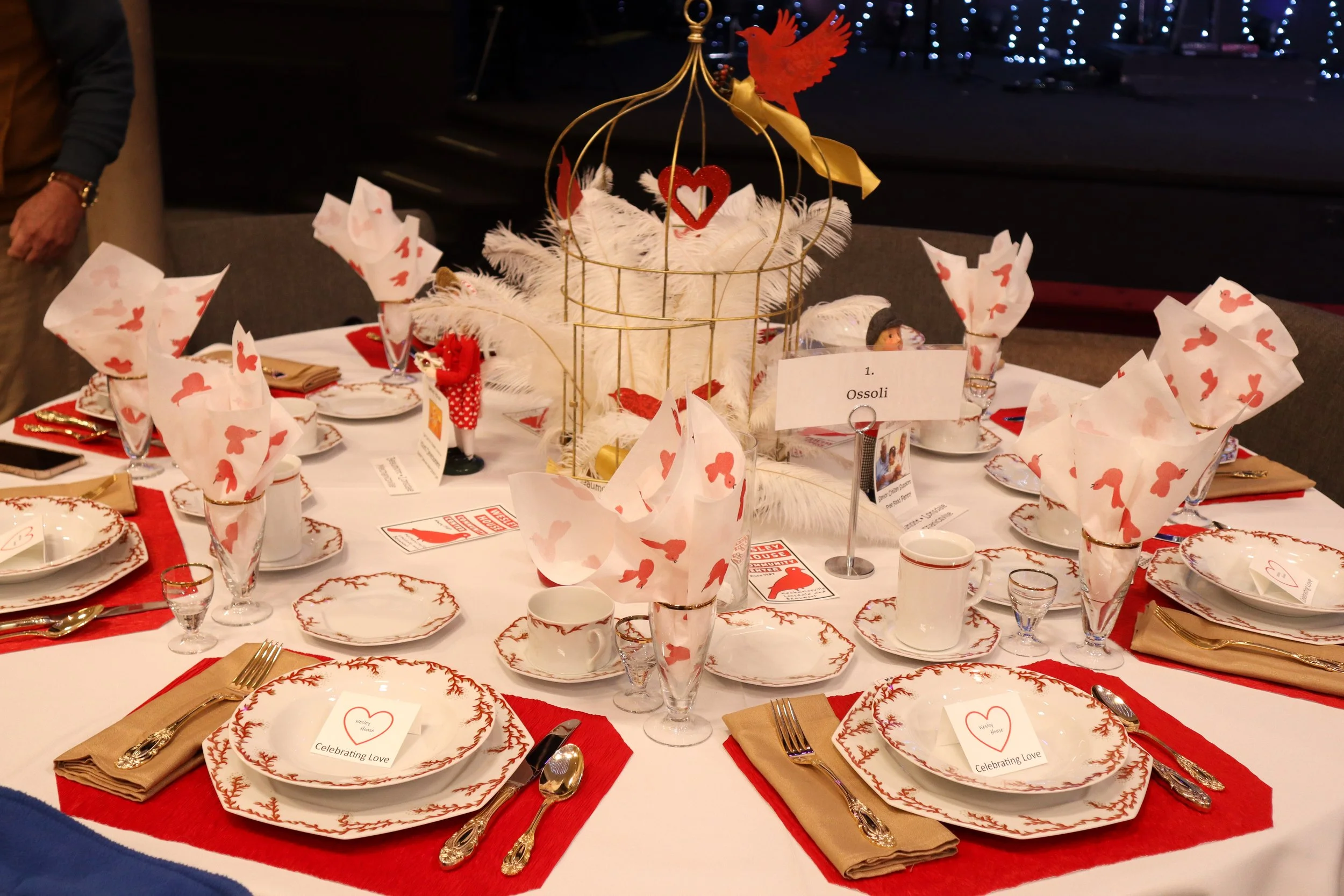 A round table set for a festive holiday dinner with red, white, and gold themed tableware and decor. There are plates with gold and red designs, gold utensils, white cups, and napkins decorated with red dog-shaped patterns. Centered on the table is a