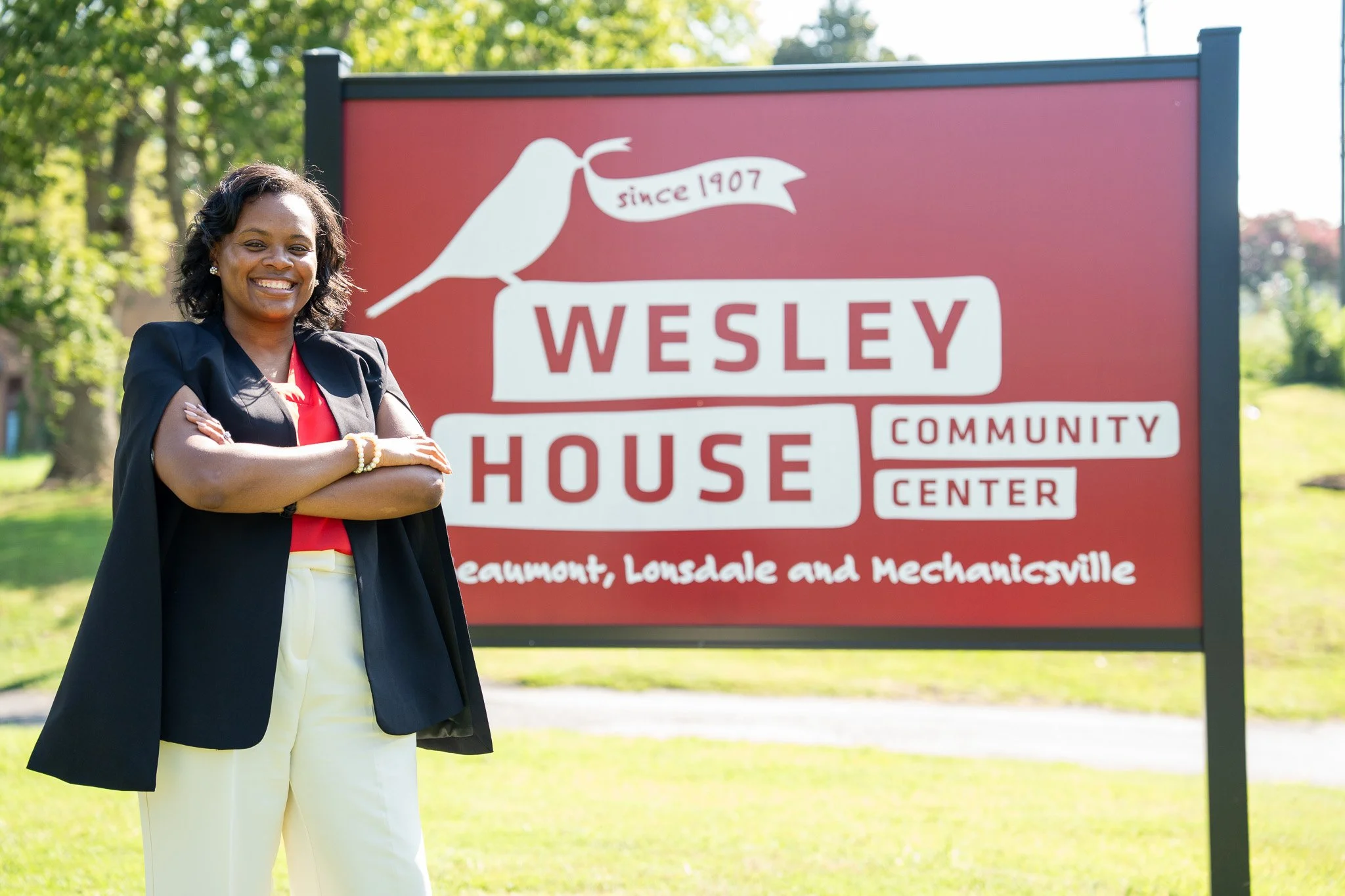 A smiling woman with arms crossed standing in front of a red sign that reads 'Wesley House Community Center, Beaumount, Lonsdale and Mechanicsville,' outdoors with trees in the background.