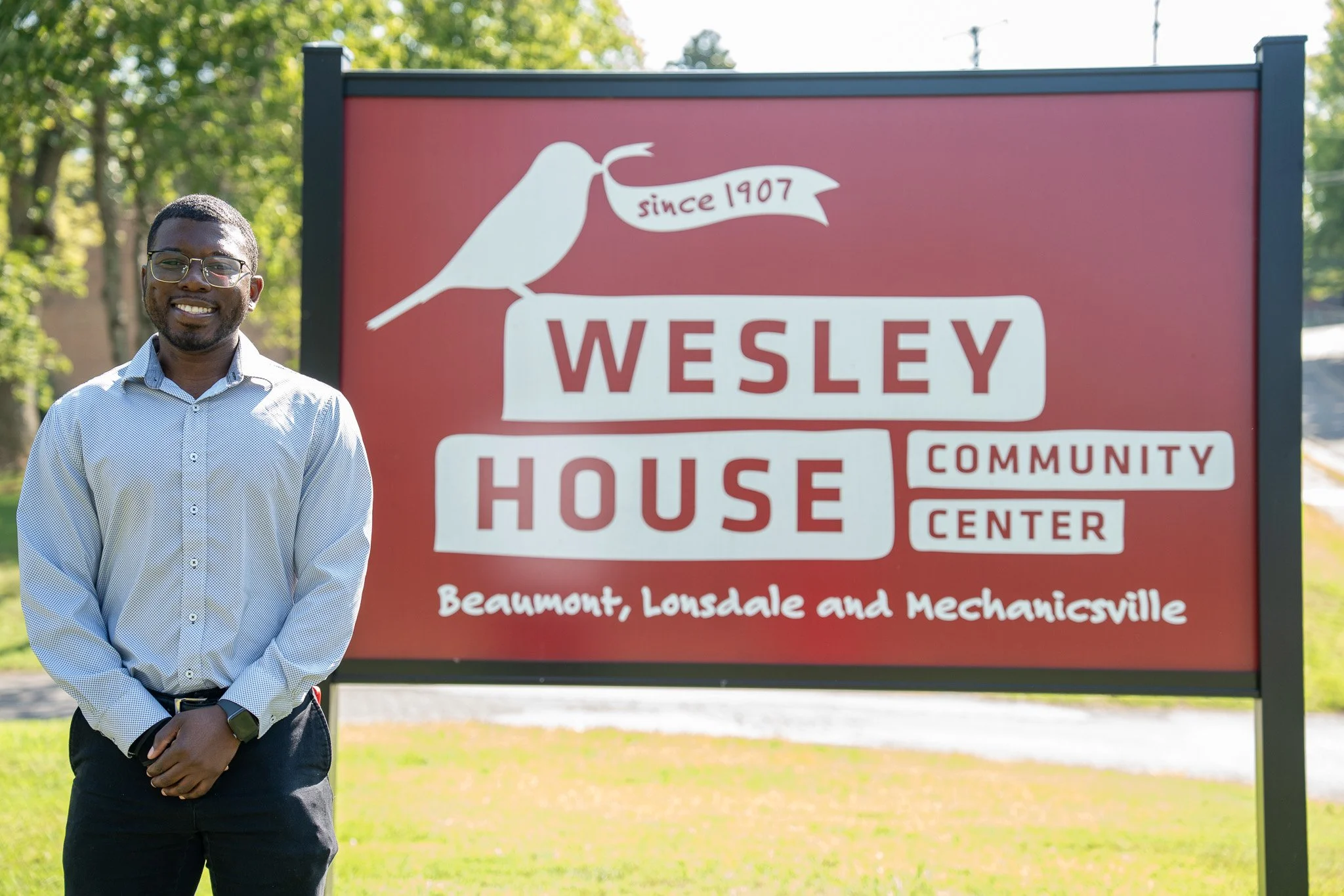 A smiling man wearing glasses, a light blue button-up shirt, and a watch, standing in front of a red sign that reads "Wesley House Community Center, Beaumont, Lousdale, and Mchenicsville." The sign features a white bird silhouette with a banner reading "since 1907."
