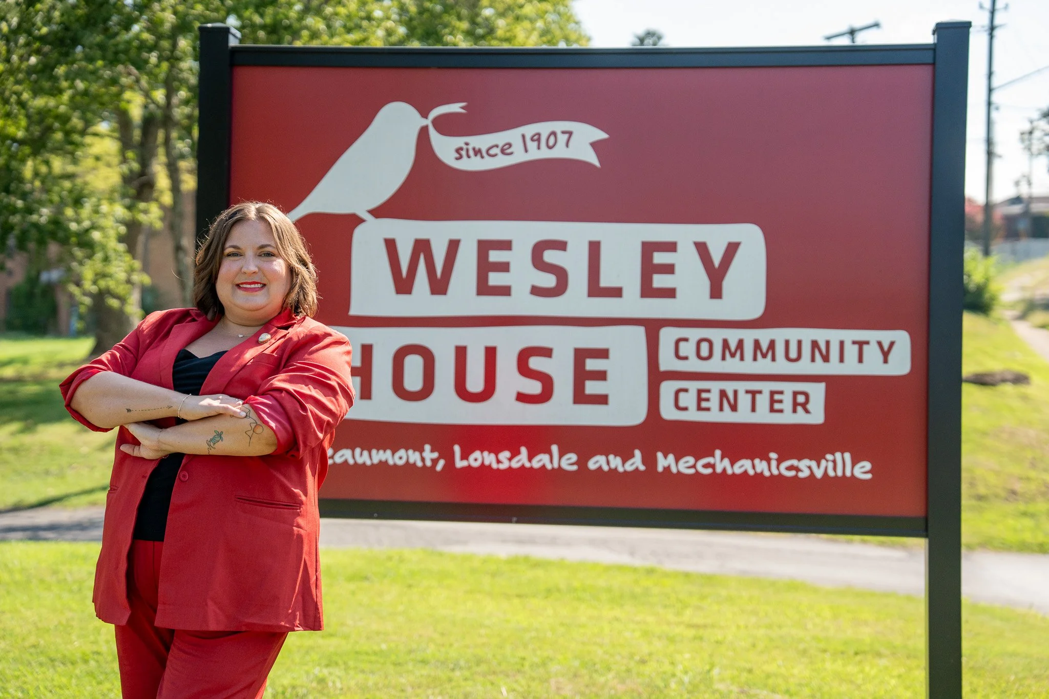 A woman in a red blazer and pants standing in front of a red sign that reads "Wesley House Community Center" with the text "since 1907" and locations Beaumont, Lonsdale, and Mechanicsville. She is smiling with her arms crossed.