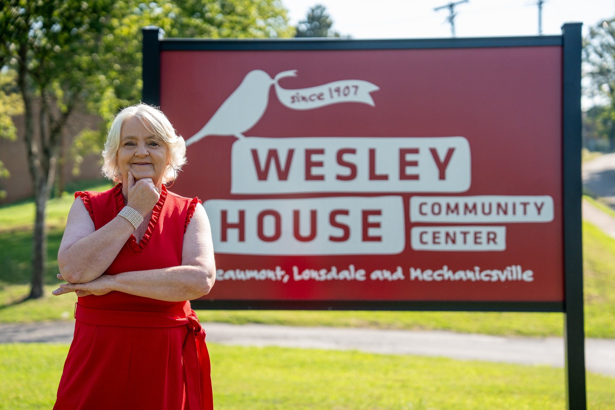An elderly woman with white hair wearing a red dress standing outdoors in front of a sign for Wesley House Community Center in Southeast Lonsdale and Mechanicsville.