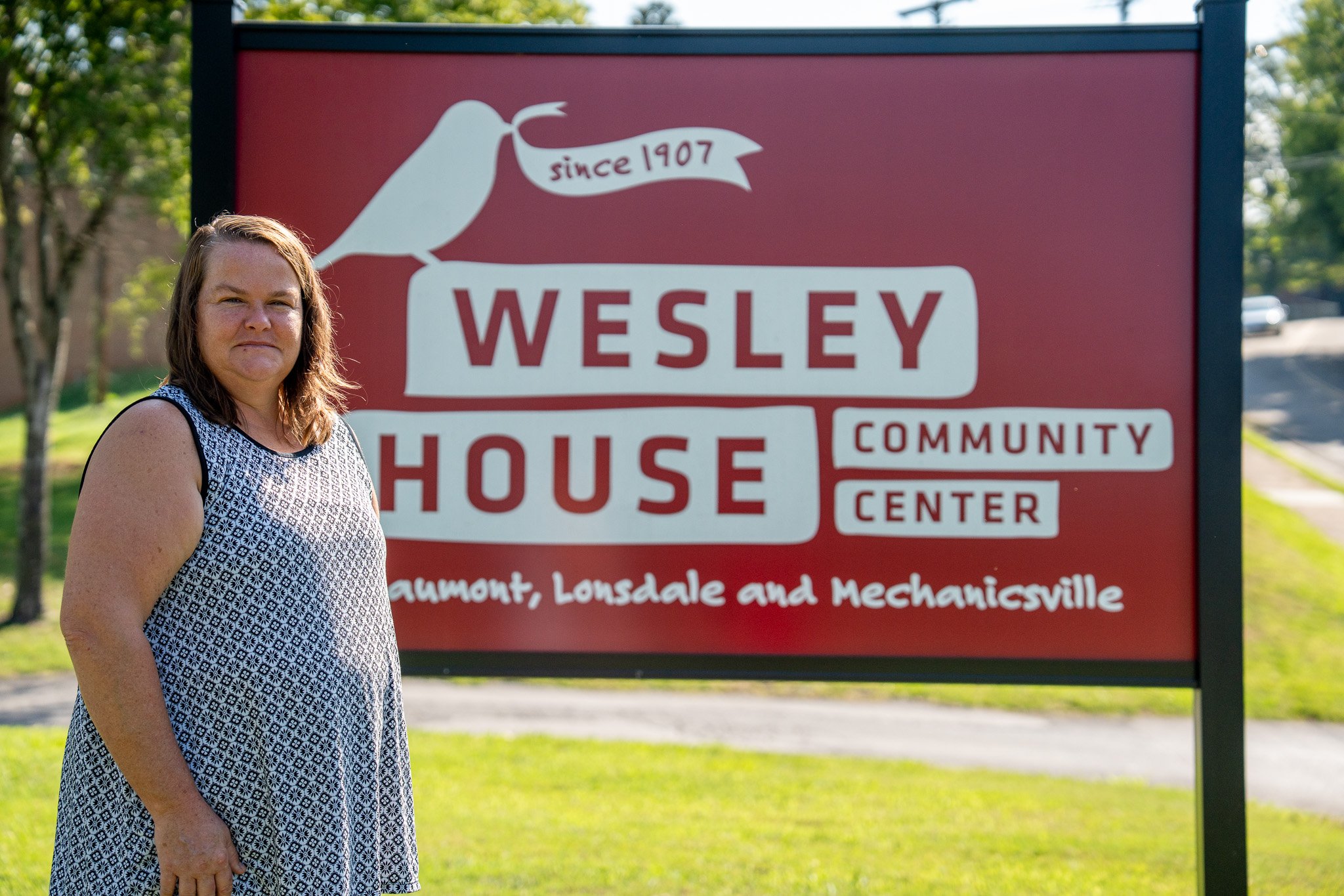 A woman standing outdoors in front of a sign that reads 'Wesley House Community Center' with locations listed as Gaumont, Lonsdale, and Mechanicsville.
