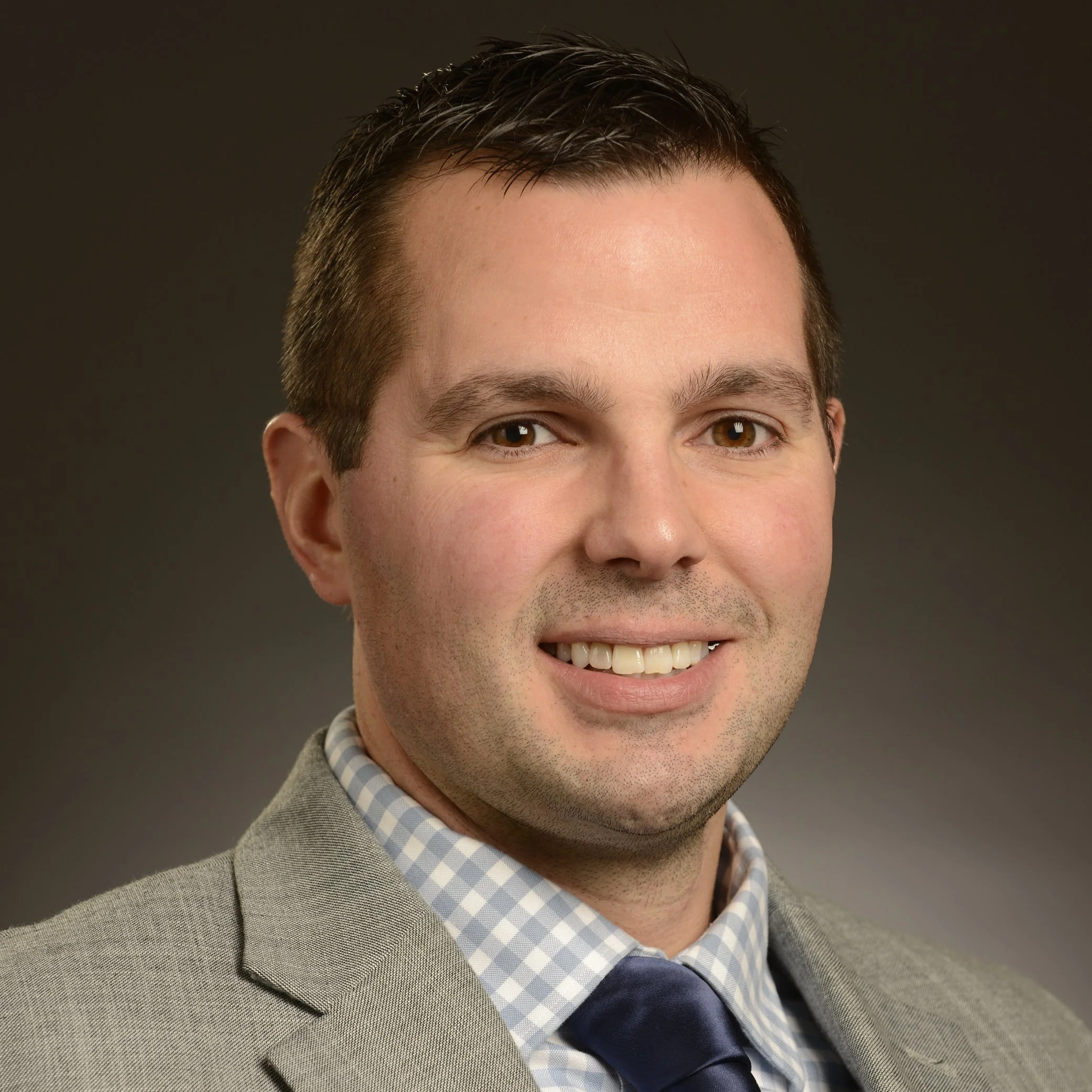 Professional headshot of a smiling man in a suit and tie against a dark background.