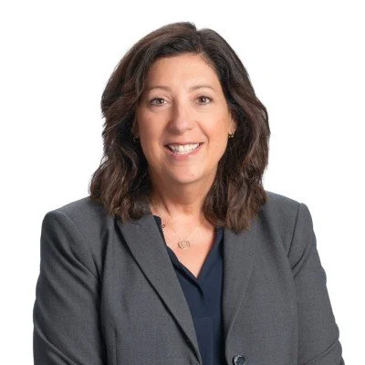 Professional woman with shoulder-length dark hair in a gray blazer smiling against a white background.